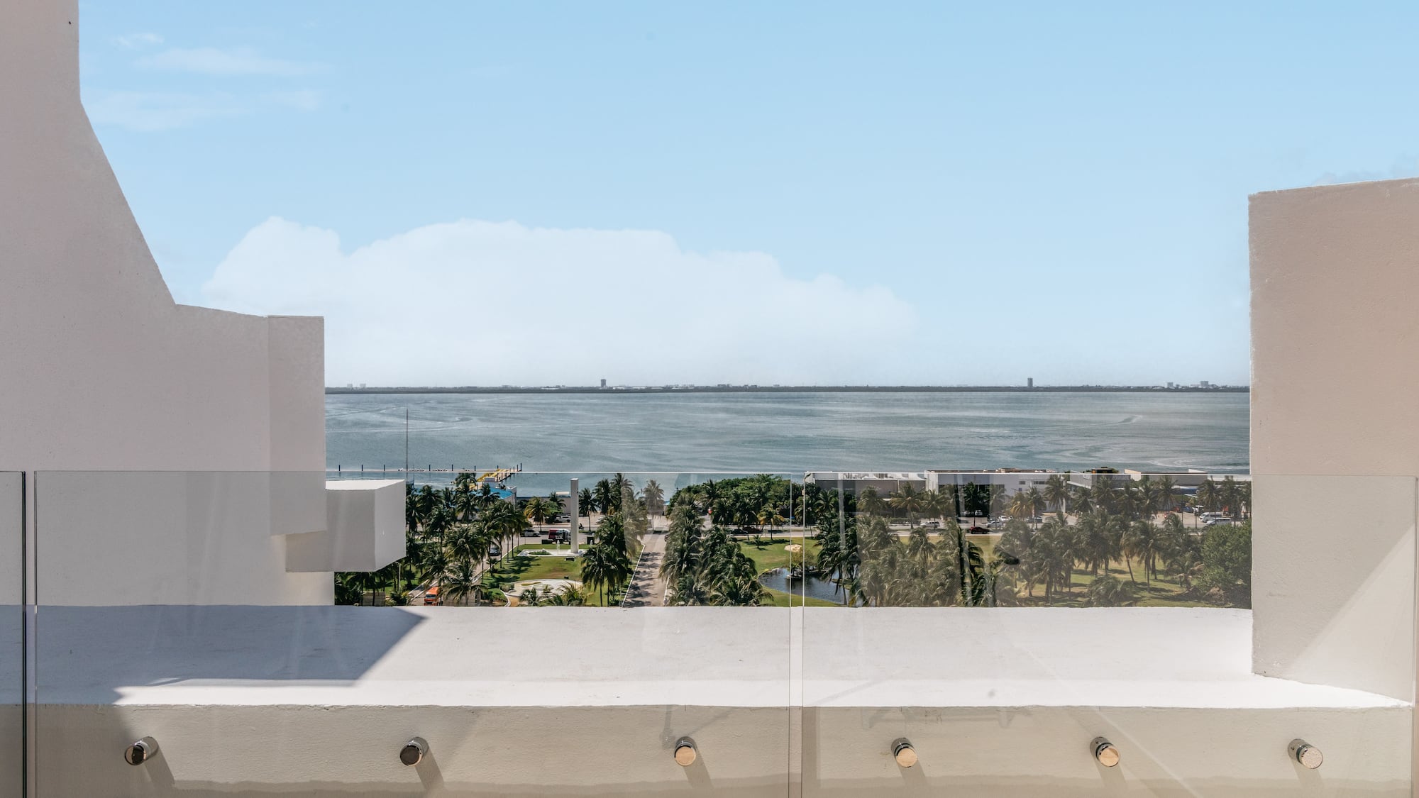 Balcony with glass railing overlooking bay, palm trees, and distant city skyline.