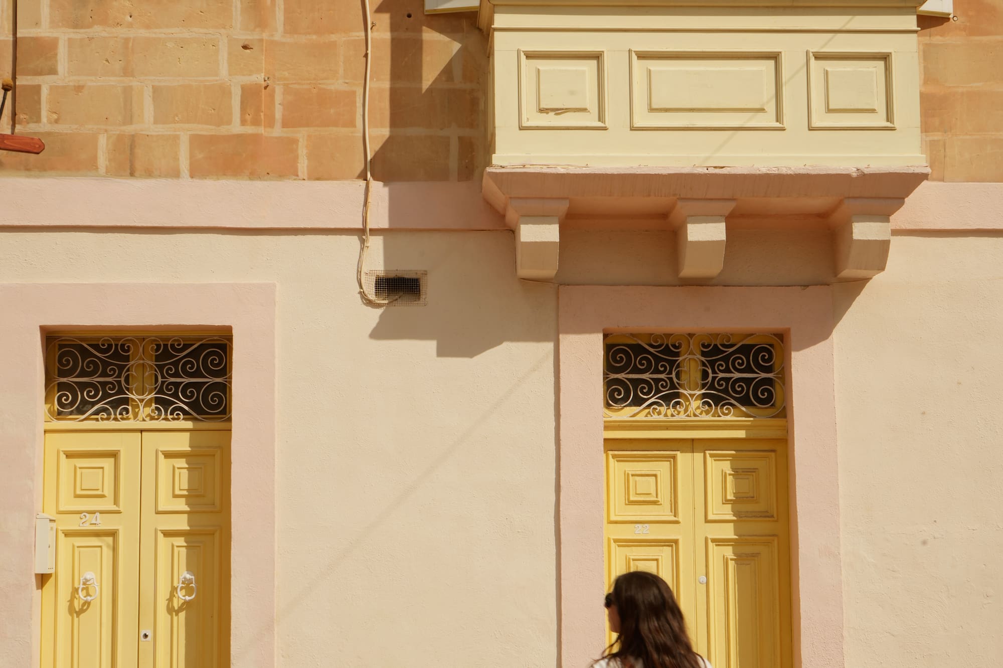 Woman walking on sunny street by yellow doors, traditional Maltese balcony.