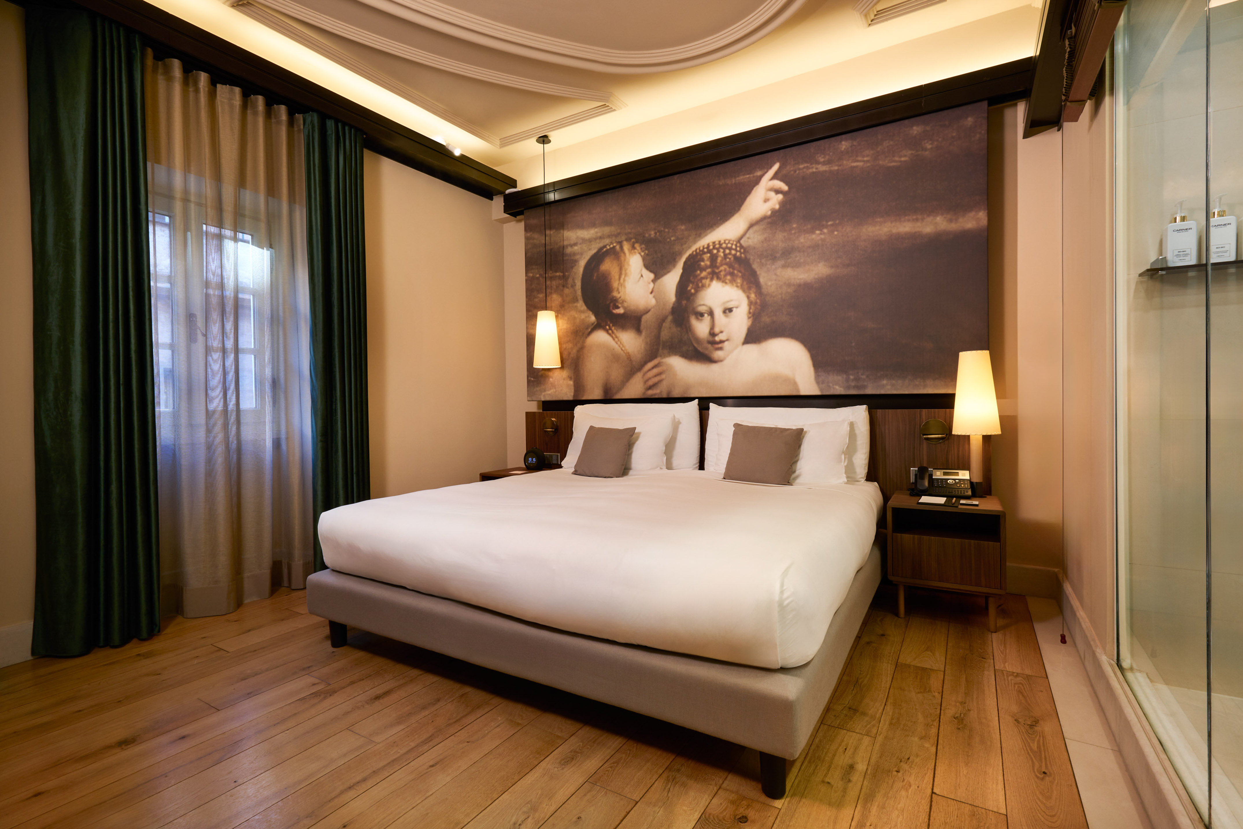 Modern hotel bedroom with large bed, renaissance art, wood floor, and green curtains.
