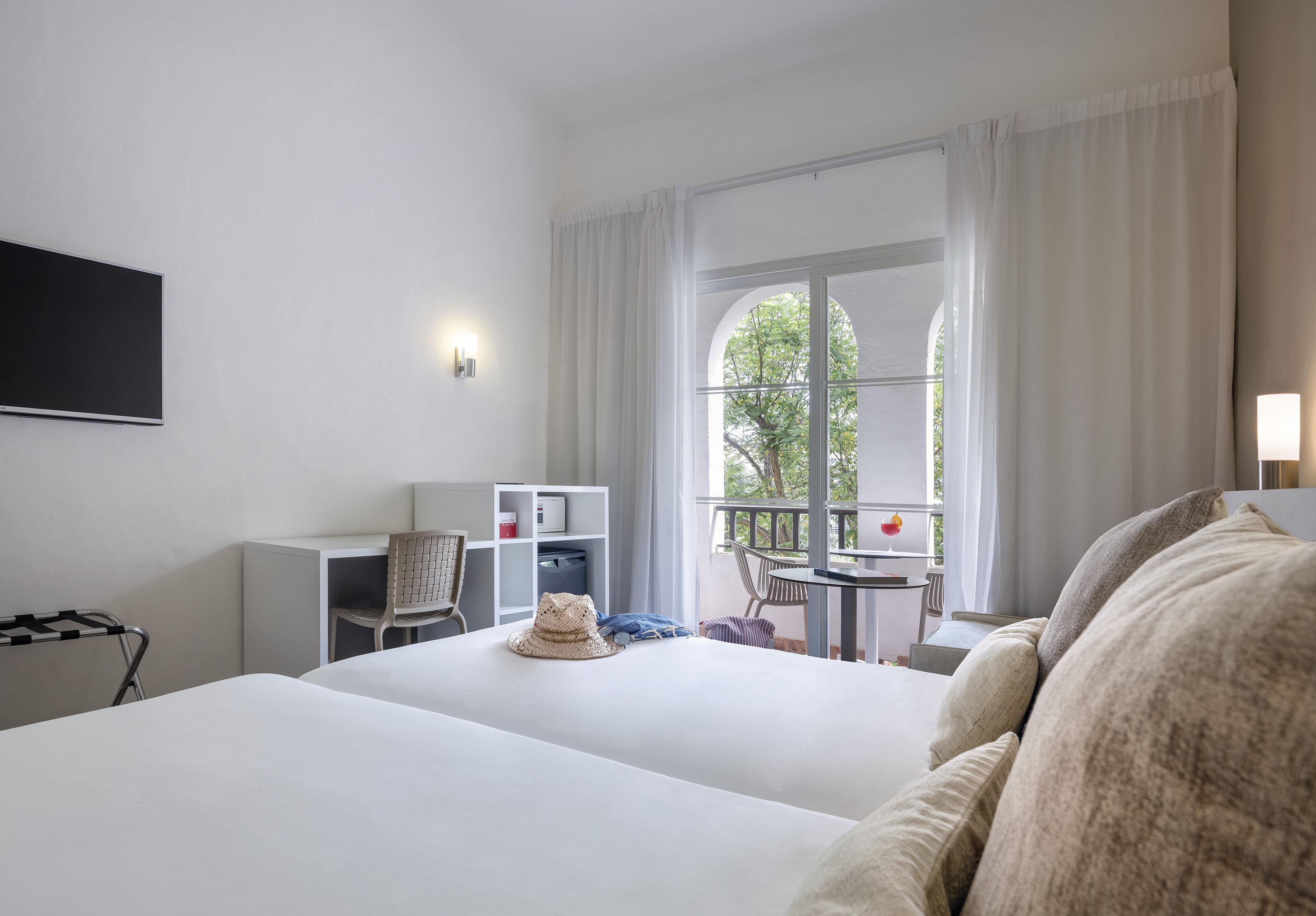 Hotel room with twin beds, desk, wall TV, and private balcony with outdoor seating.