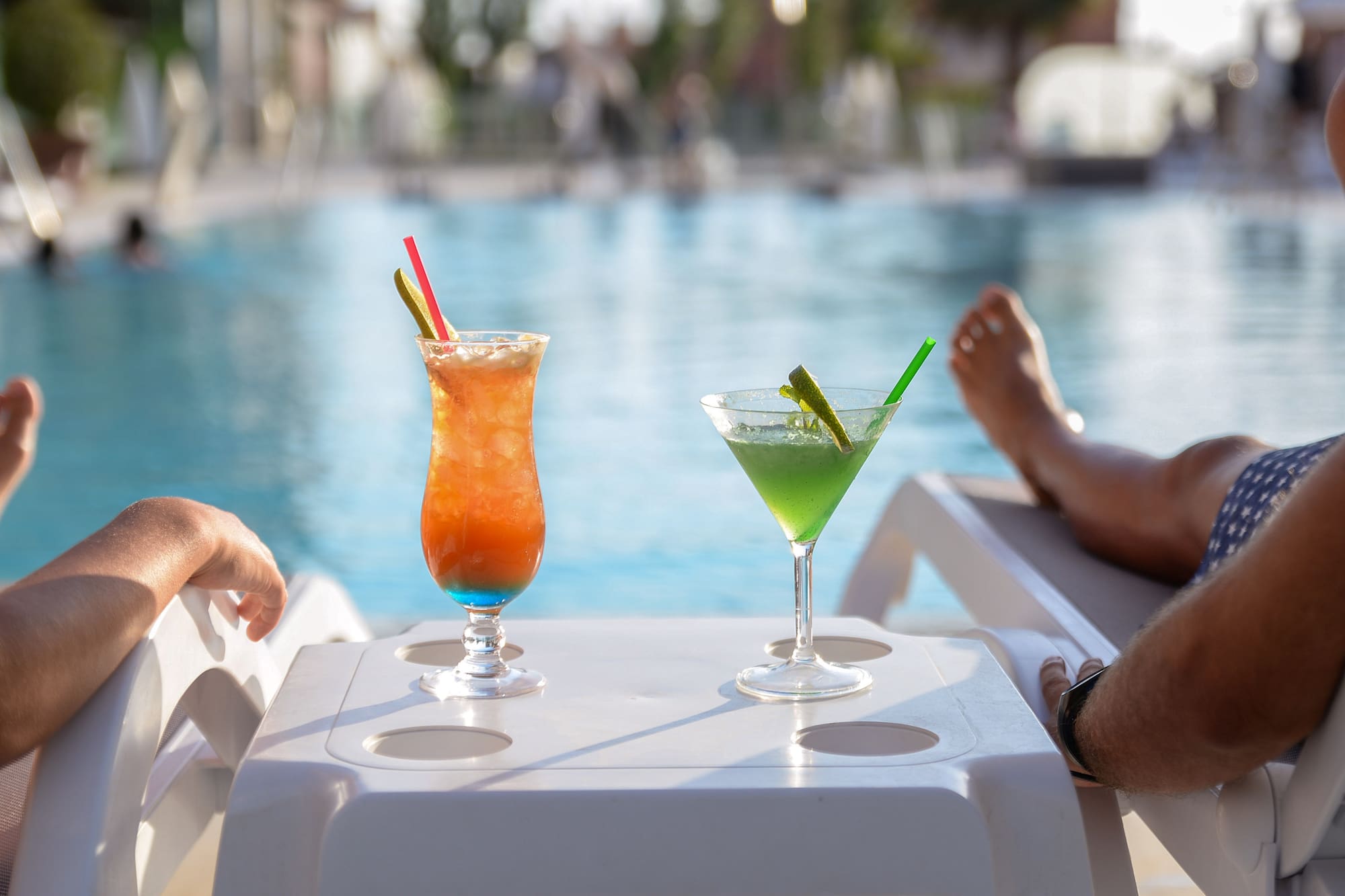 Two colorful cocktails by the pool, people relaxing, summer vacation.