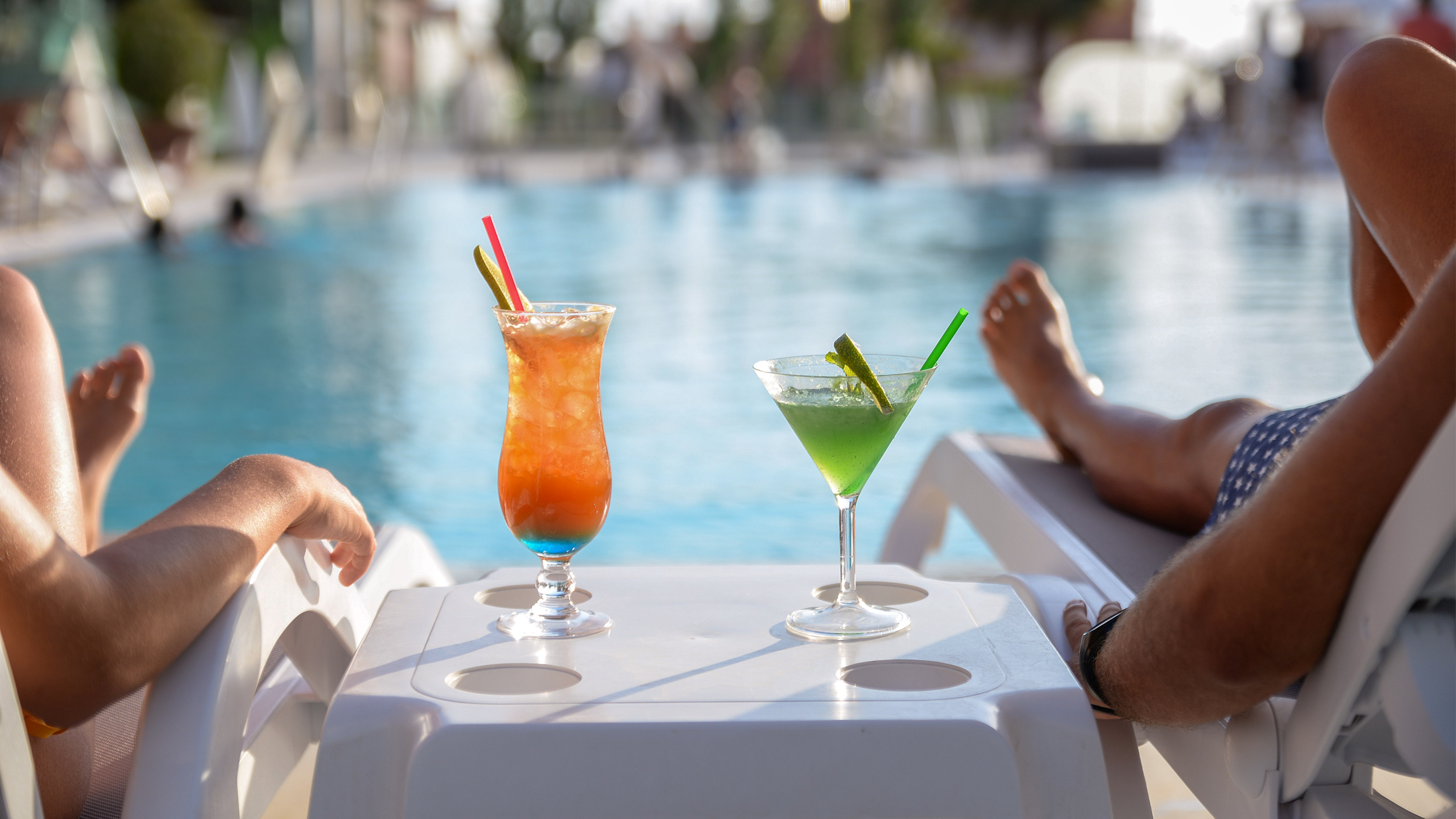 Two colorful cocktails by the pool, people relaxing, summer vacation.