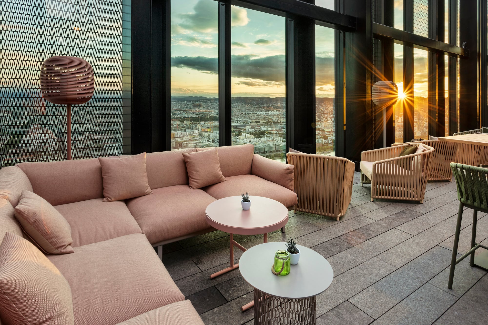 Rooftop lounge with pink sofa, woven chairs, and city view at sunset.