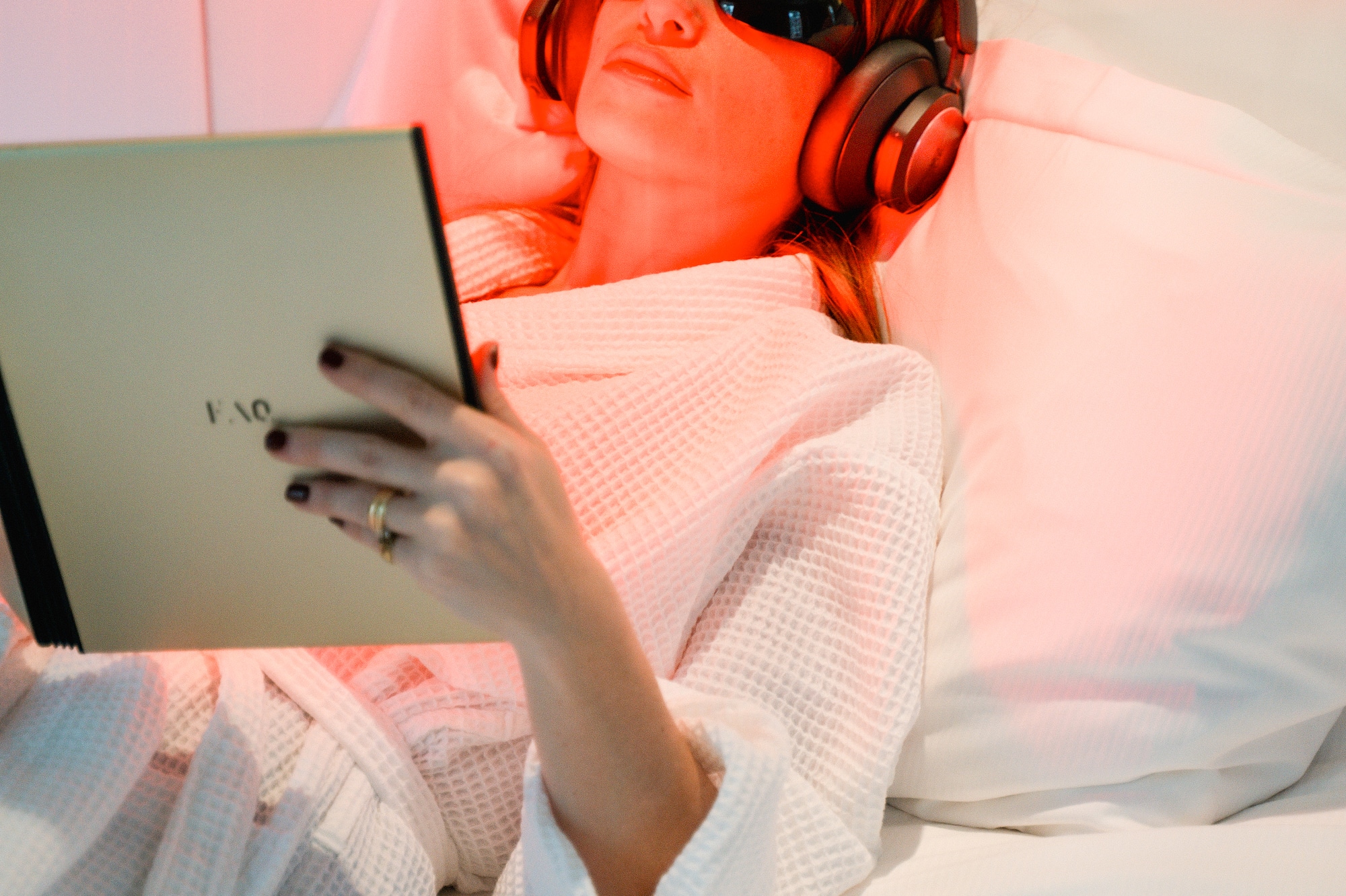 Woman in bed with red light therapy, headphones, eye protection, reading tablet.
