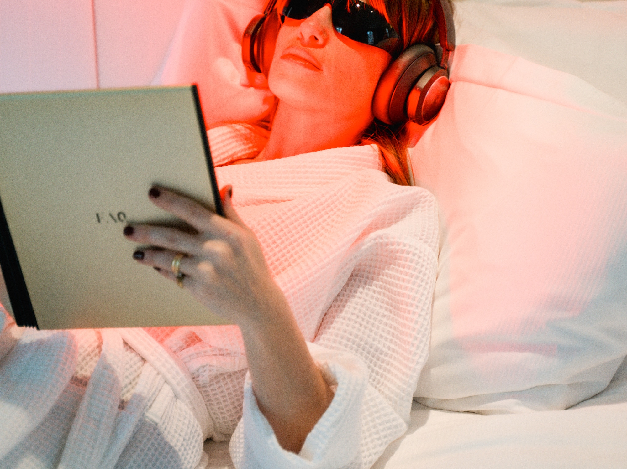Woman in bed with red light therapy, headphones, eye protection, reading tablet.
