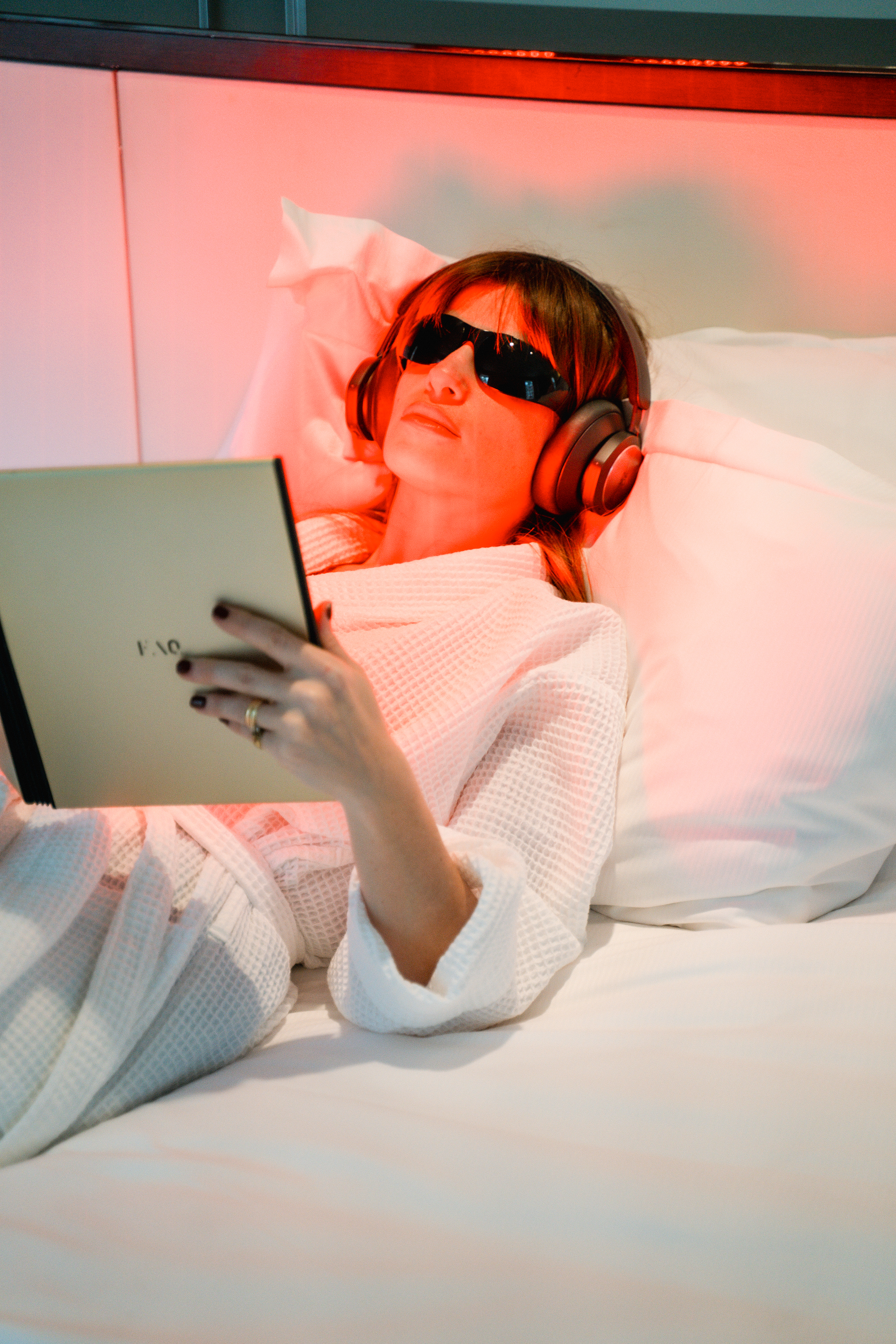 Woman in bed with red light therapy, headphones, eye protection, reading tablet.