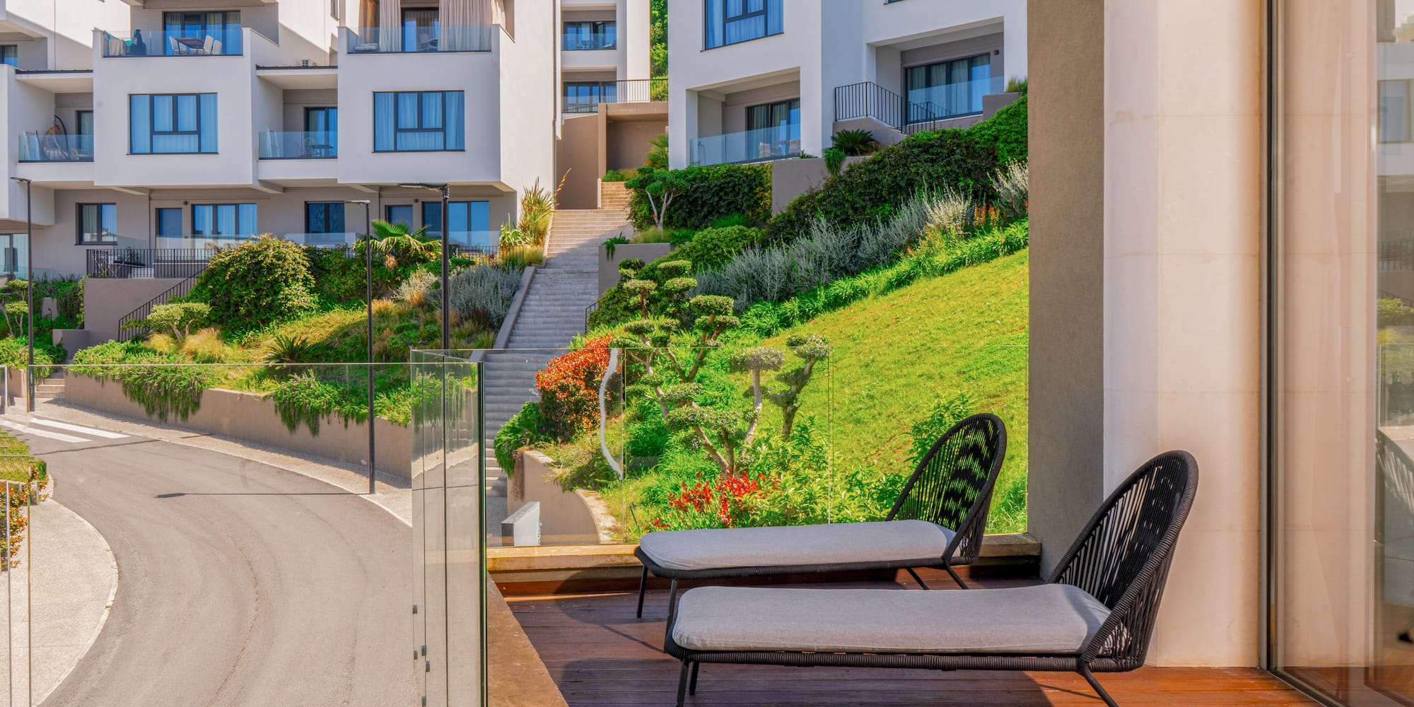 Balcony with lounge chairs, modern white buildings on a lush green hillside.
