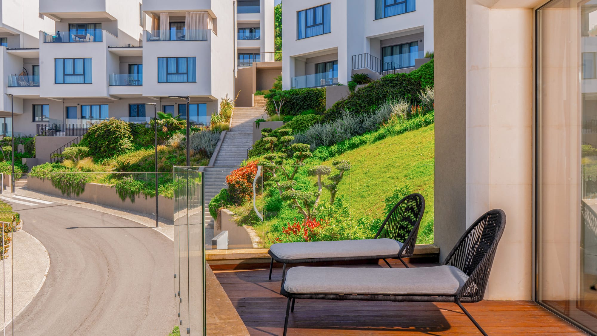 Balcony with lounge chairs, modern white buildings on a lush green hillside.