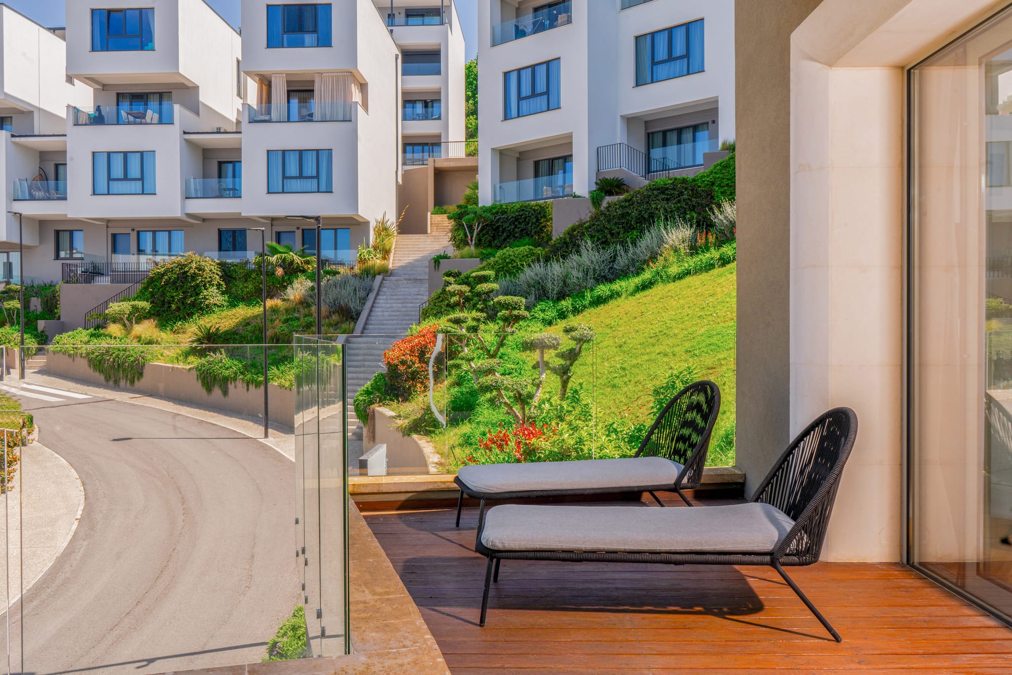 Balcony with lounge chairs, modern white buildings on a lush green hillside.