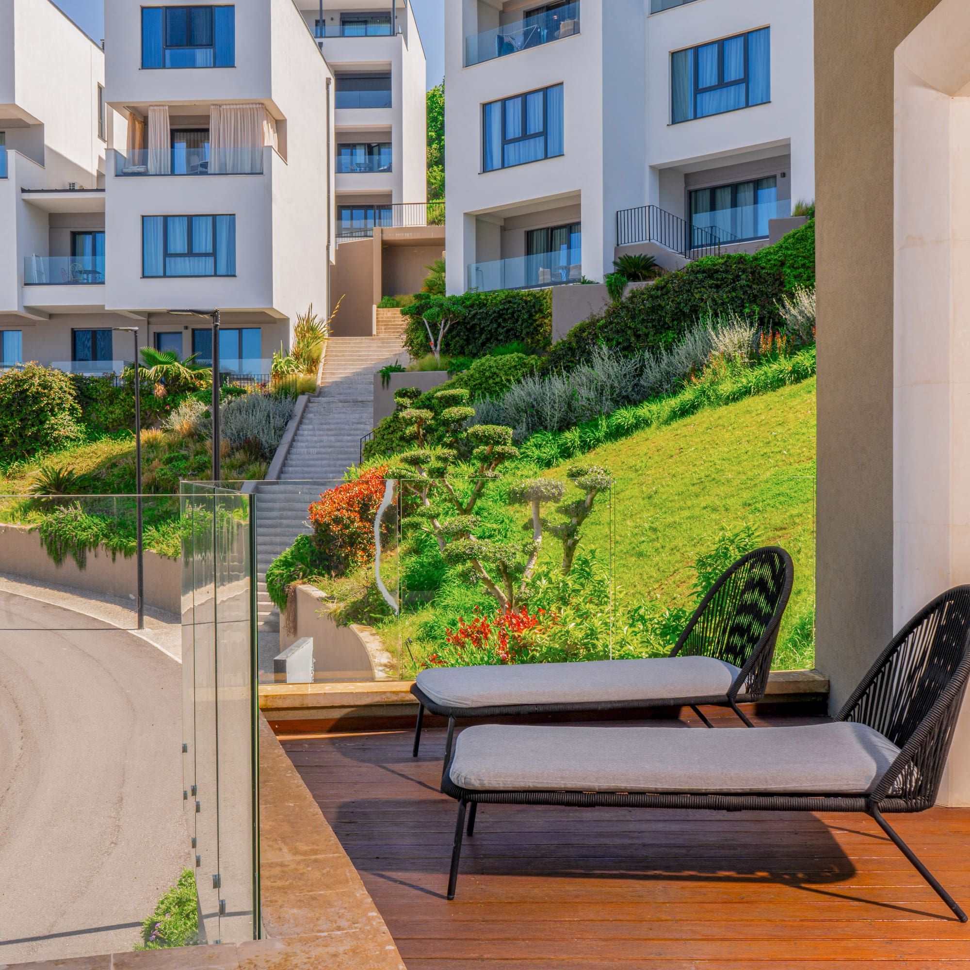 Balcony with lounge chairs, modern white buildings on a lush green hillside.