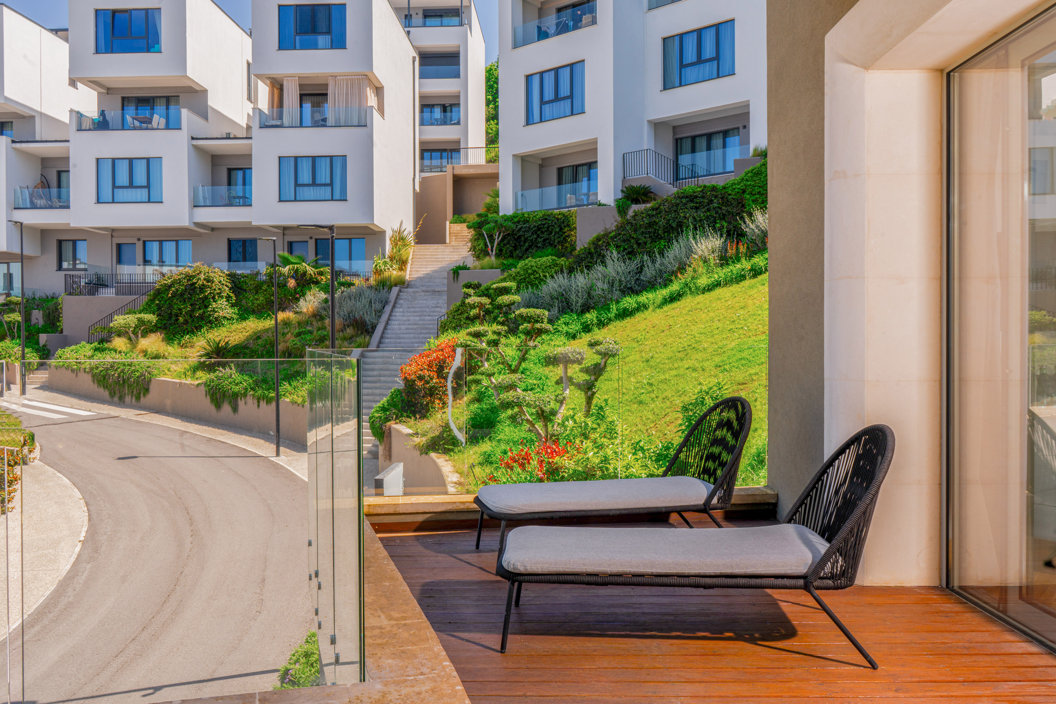 Balcony with lounge chairs, modern white buildings on a lush green hillside.
