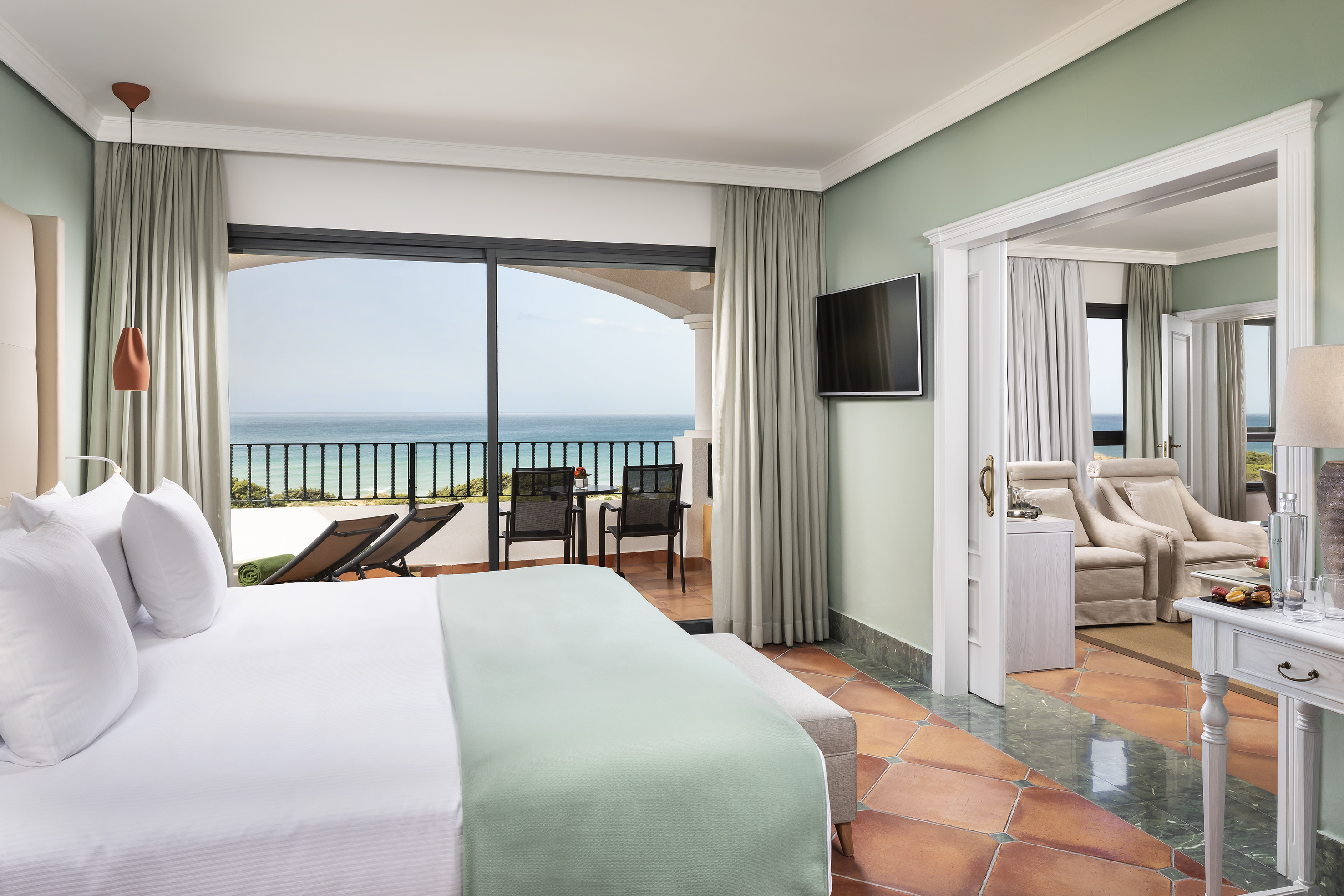 Luxury hotel room with ocean view balcony, sunbeds, and adjoining suite.