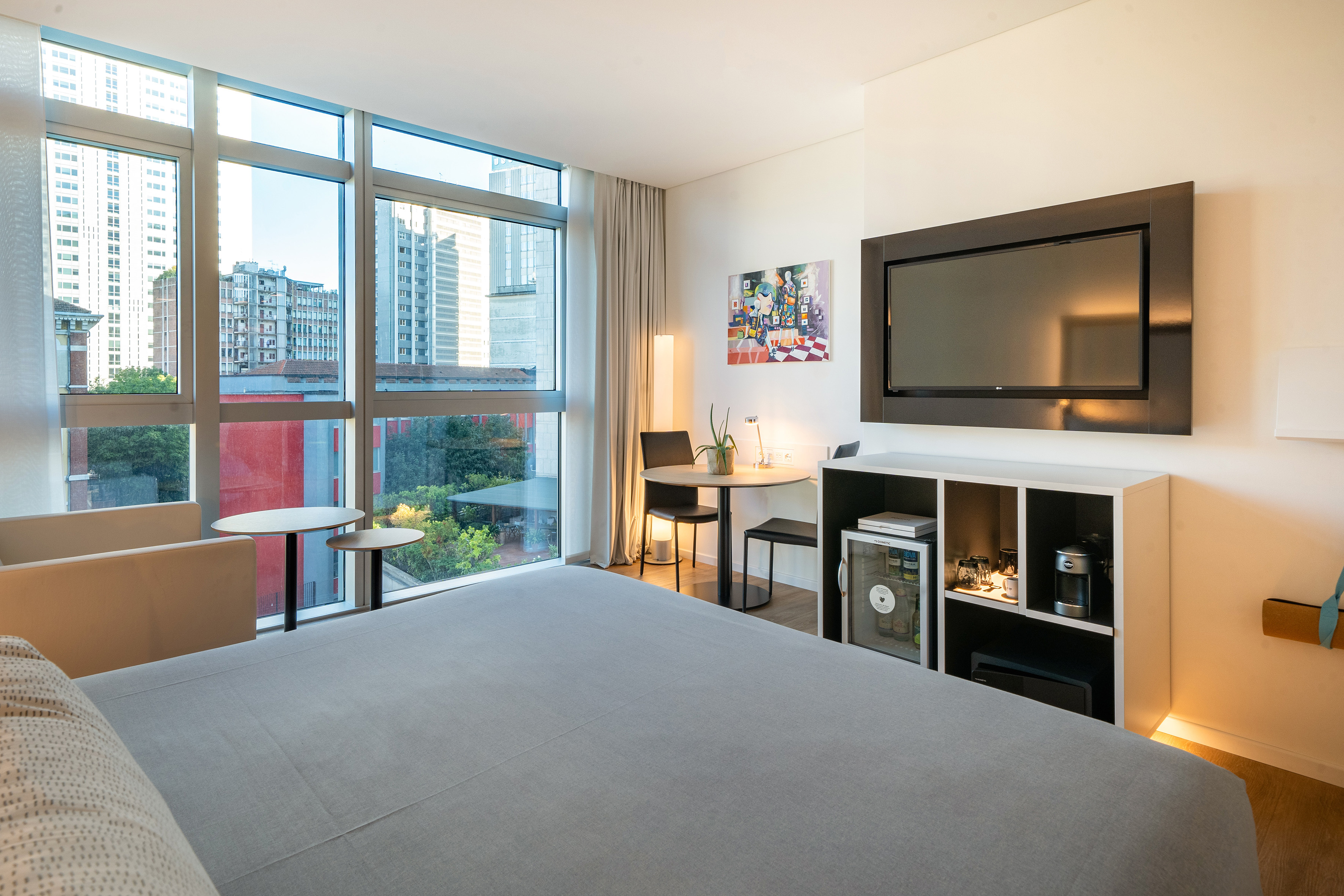 Modern hotel room with large windows, city view, bed, TV, and minibar.