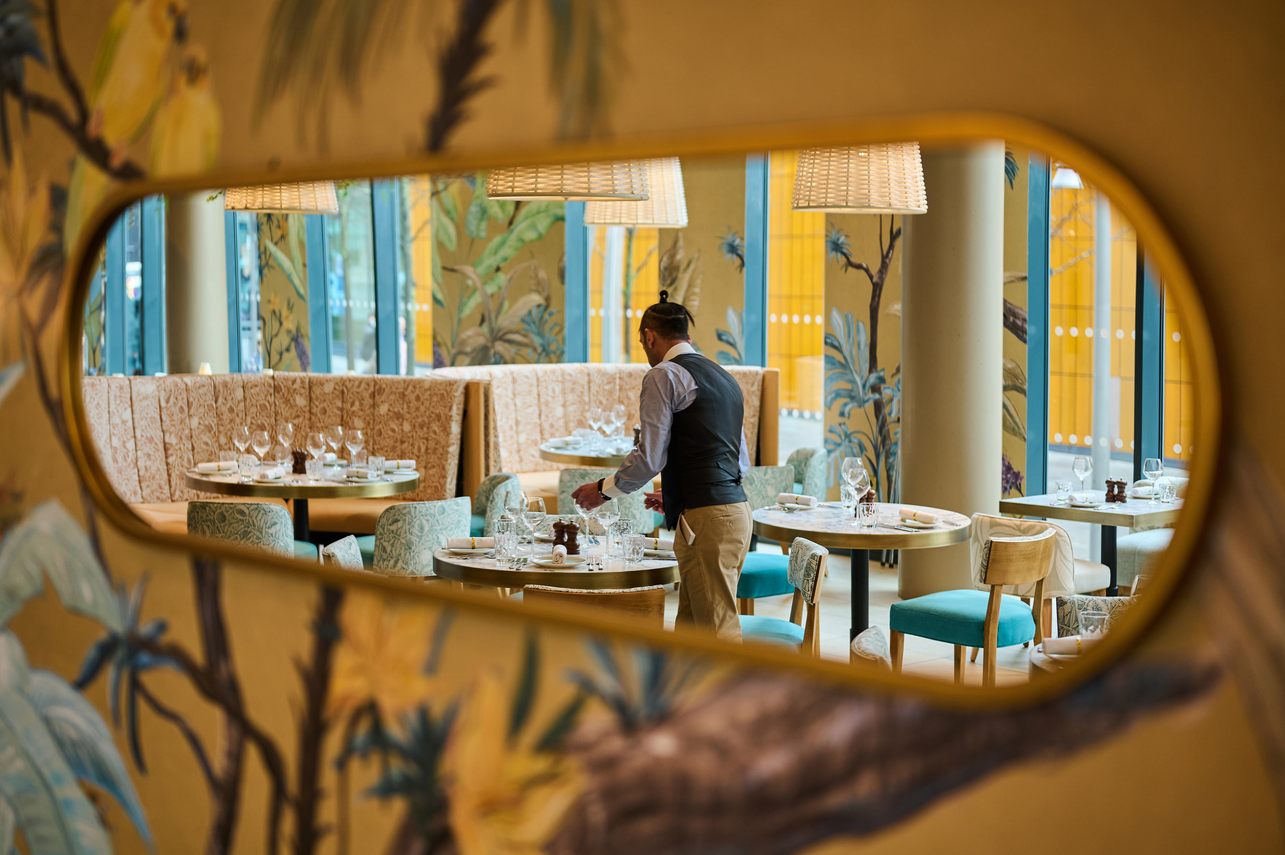Server setting tables in a tropical restaurant, reflected in a decorative mirror.