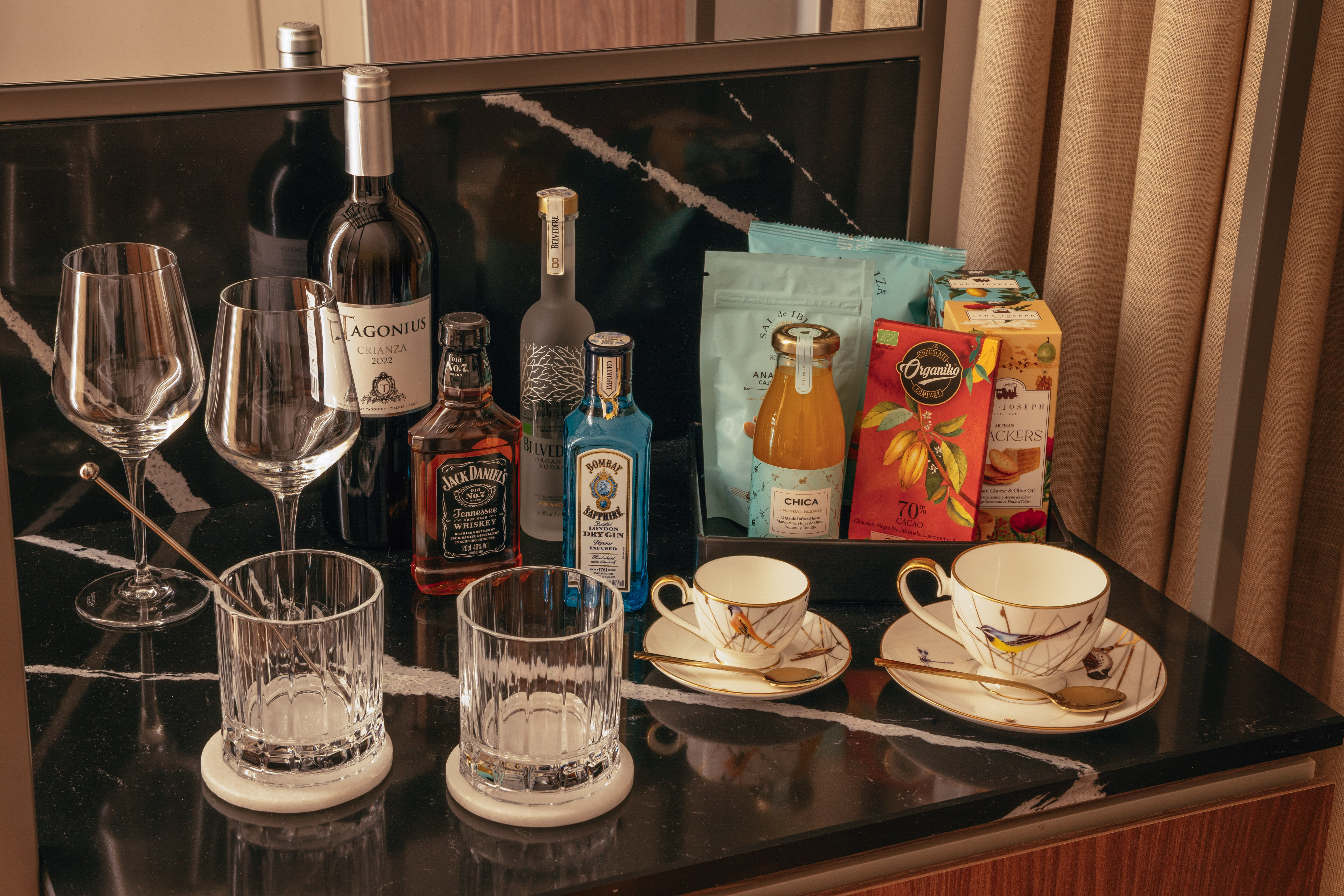 Hotel minibar with wine, spirits, glasses, gourmet snacks, chocolate, and juice.