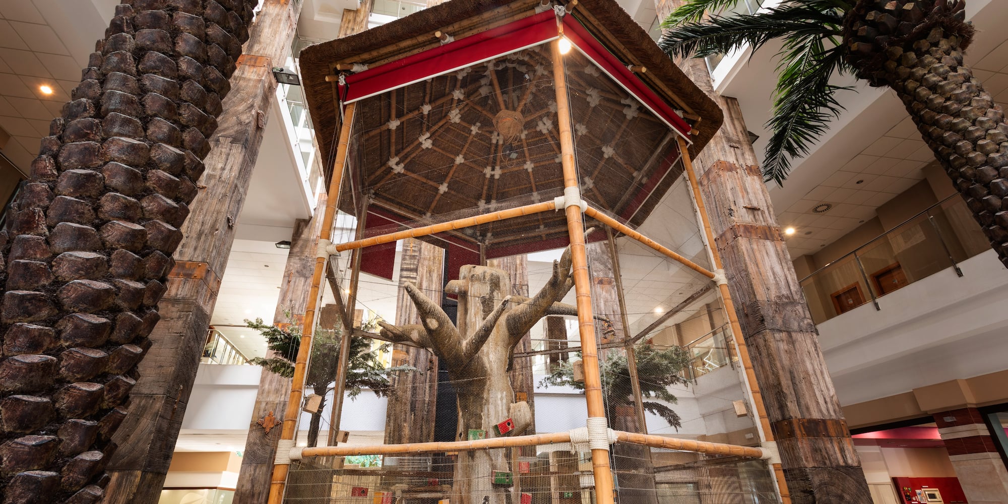 Indoor hotel aviary with thatched roof, artificial tree, and palm trees.