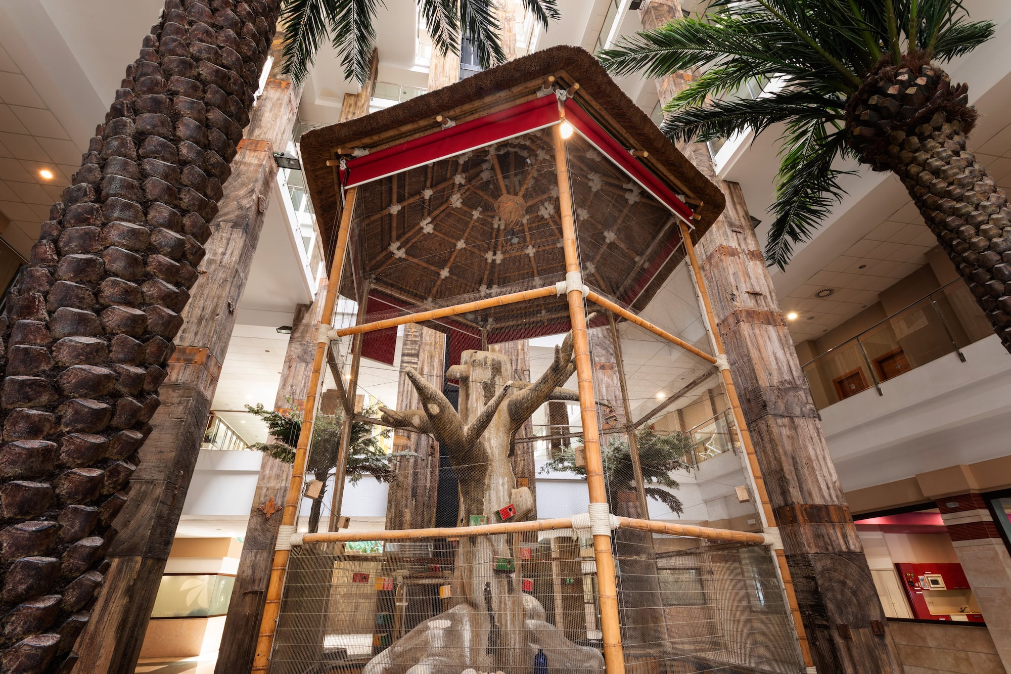 Indoor hotel aviary with thatched roof, artificial tree, and palm trees.