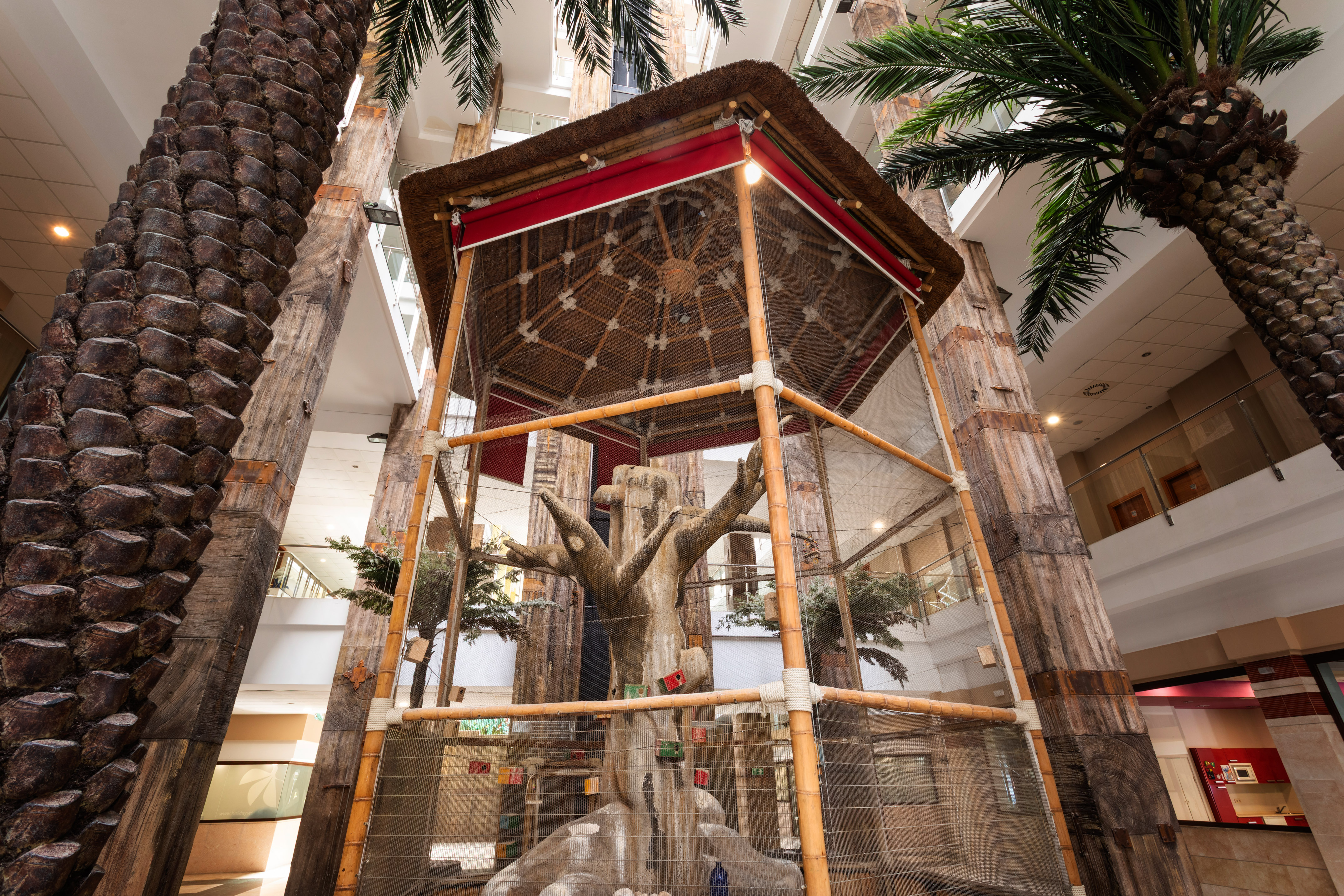 Indoor hotel aviary with thatched roof, artificial tree, and palm trees.