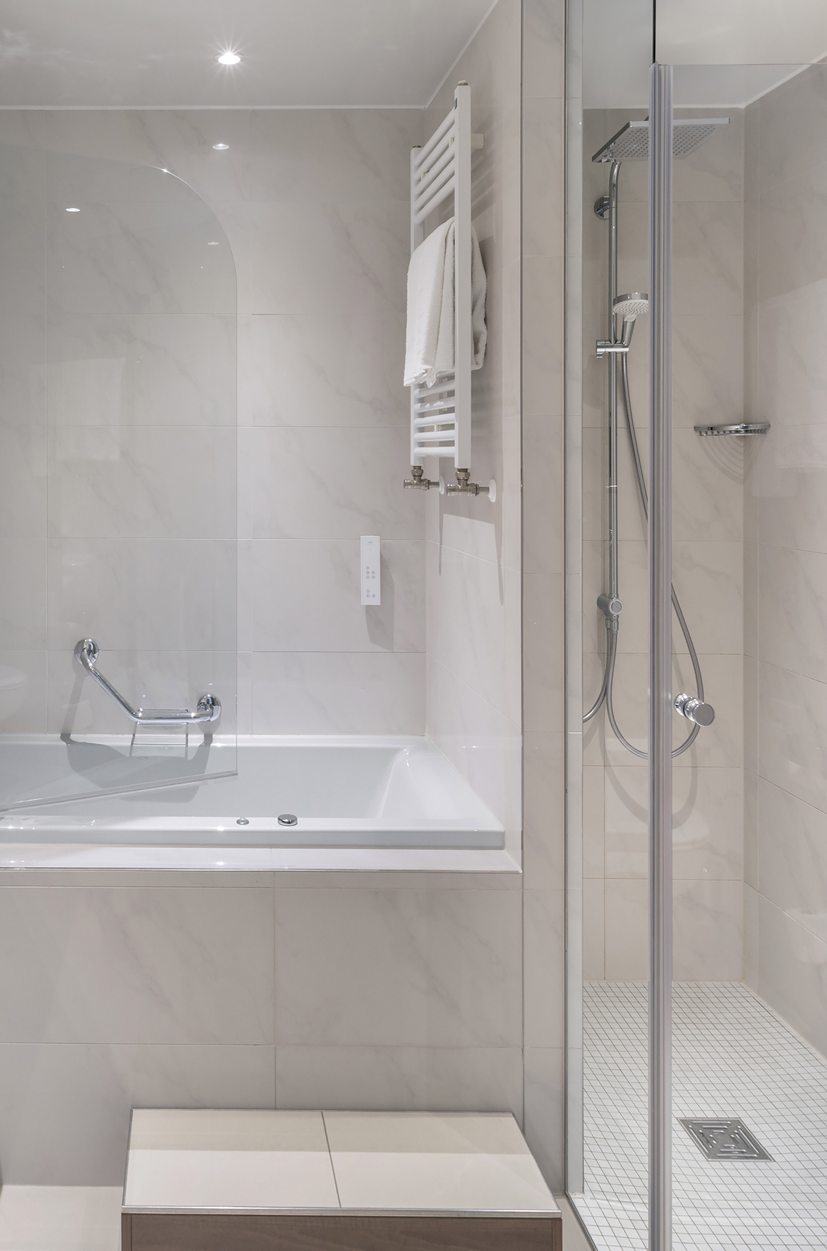 Modern white tiled bathroom with bathtub, glass shower, and towel warmer.