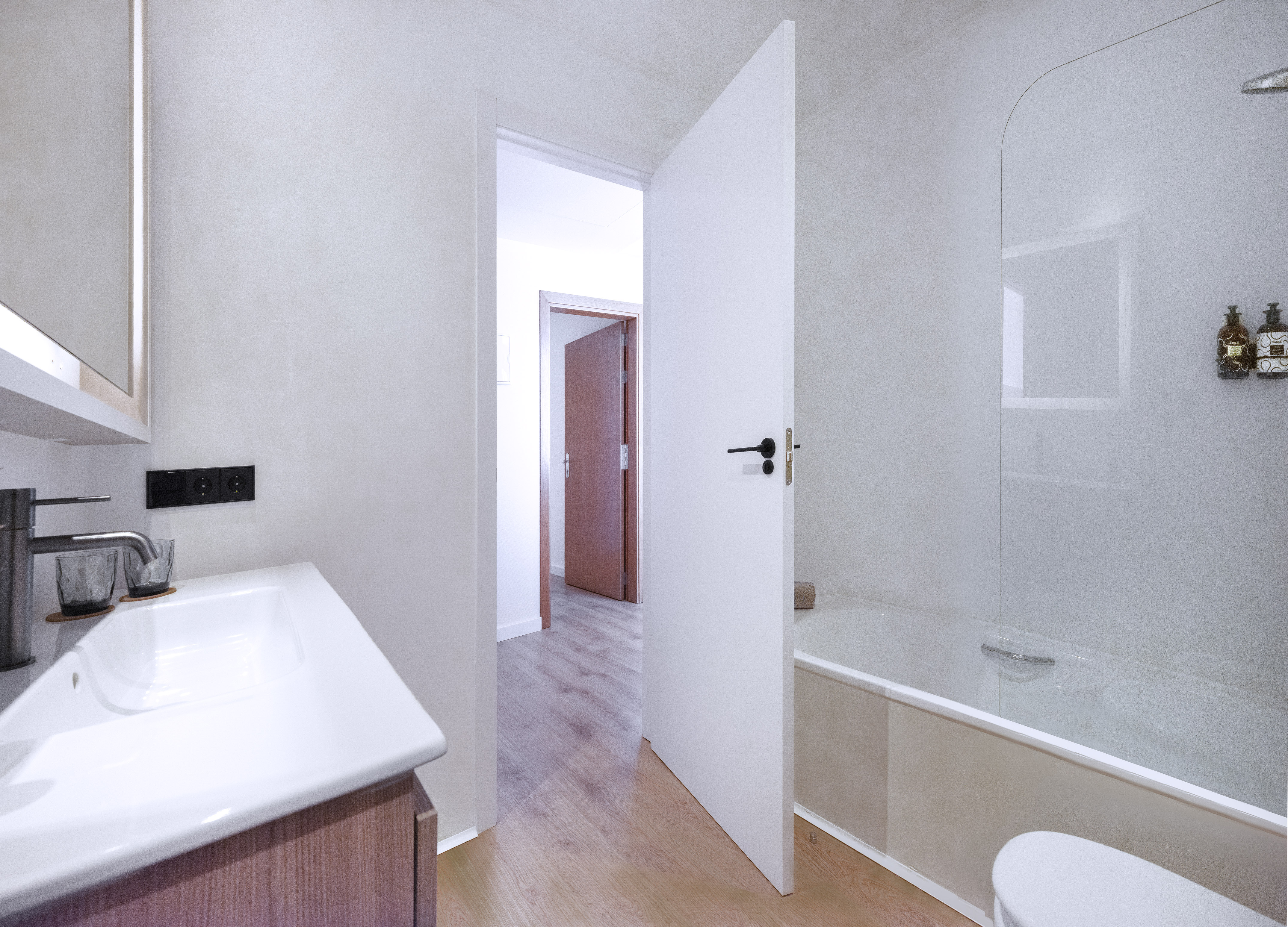 Modern white bathroom with sink, bathtub, light wood floor, and open doorway.