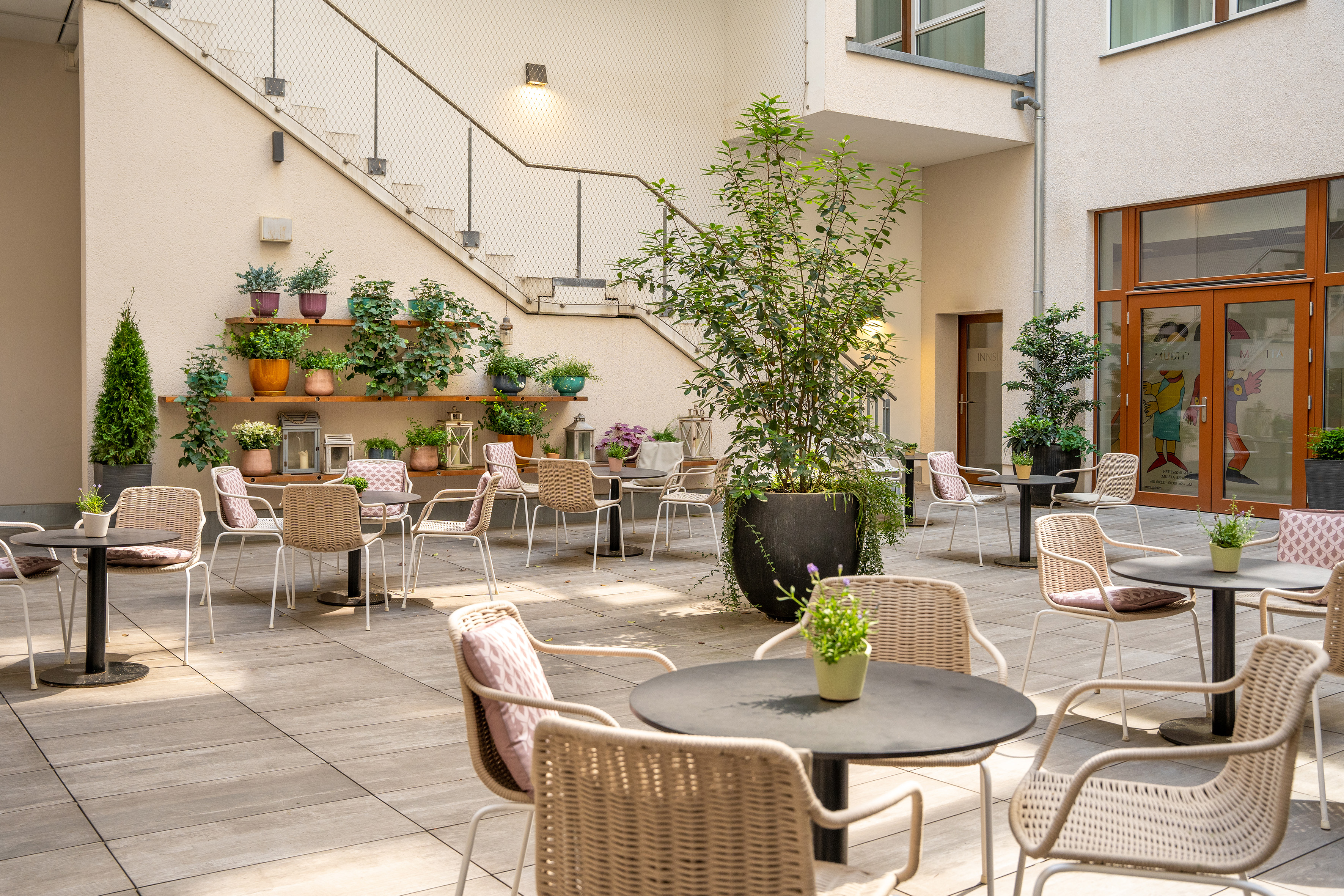 Outdoor cafe courtyard with wicker seating, black tables, and lush potted plants.