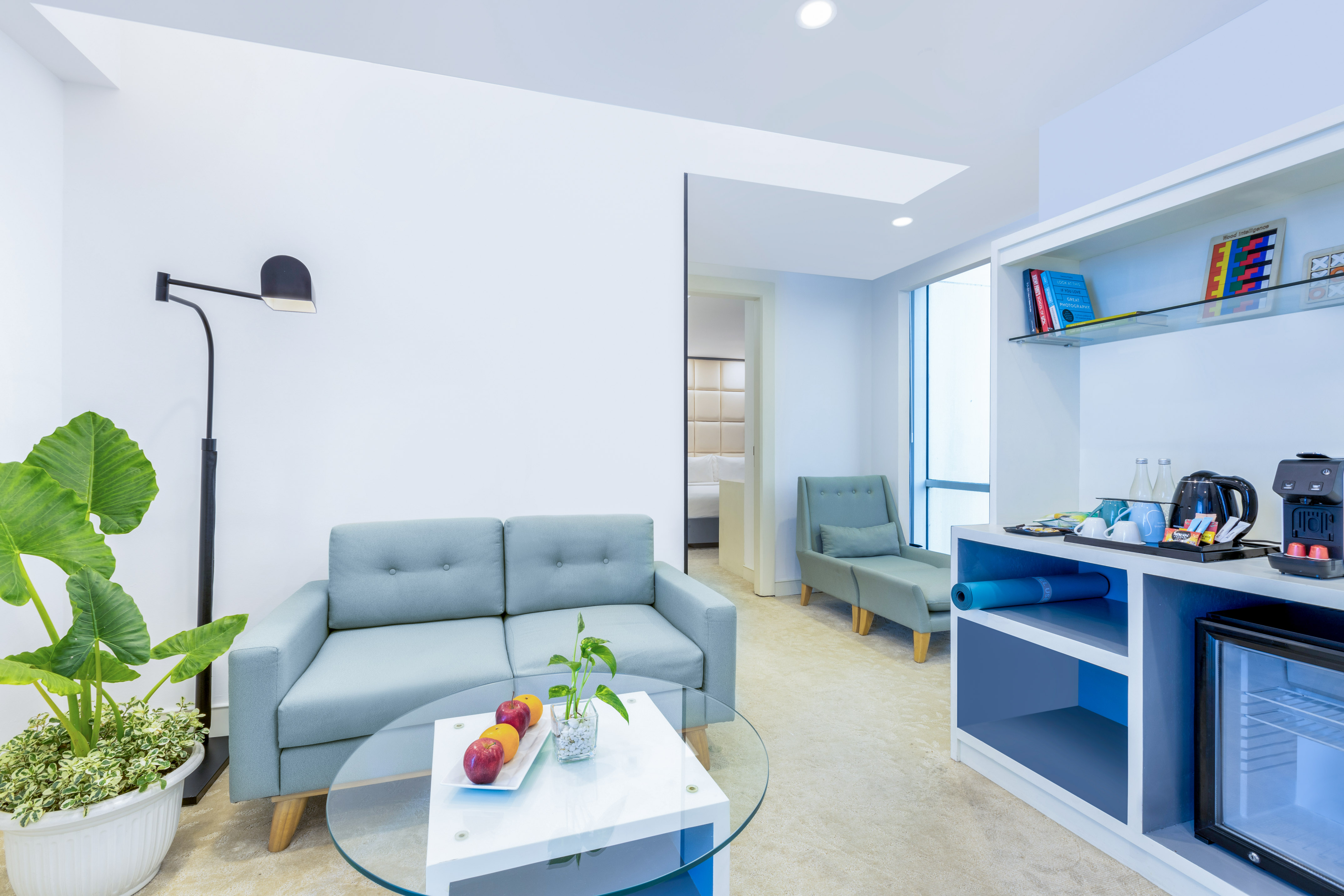 Modern hotel suite living area with light blue sofa, coffee table, mini-fridge.