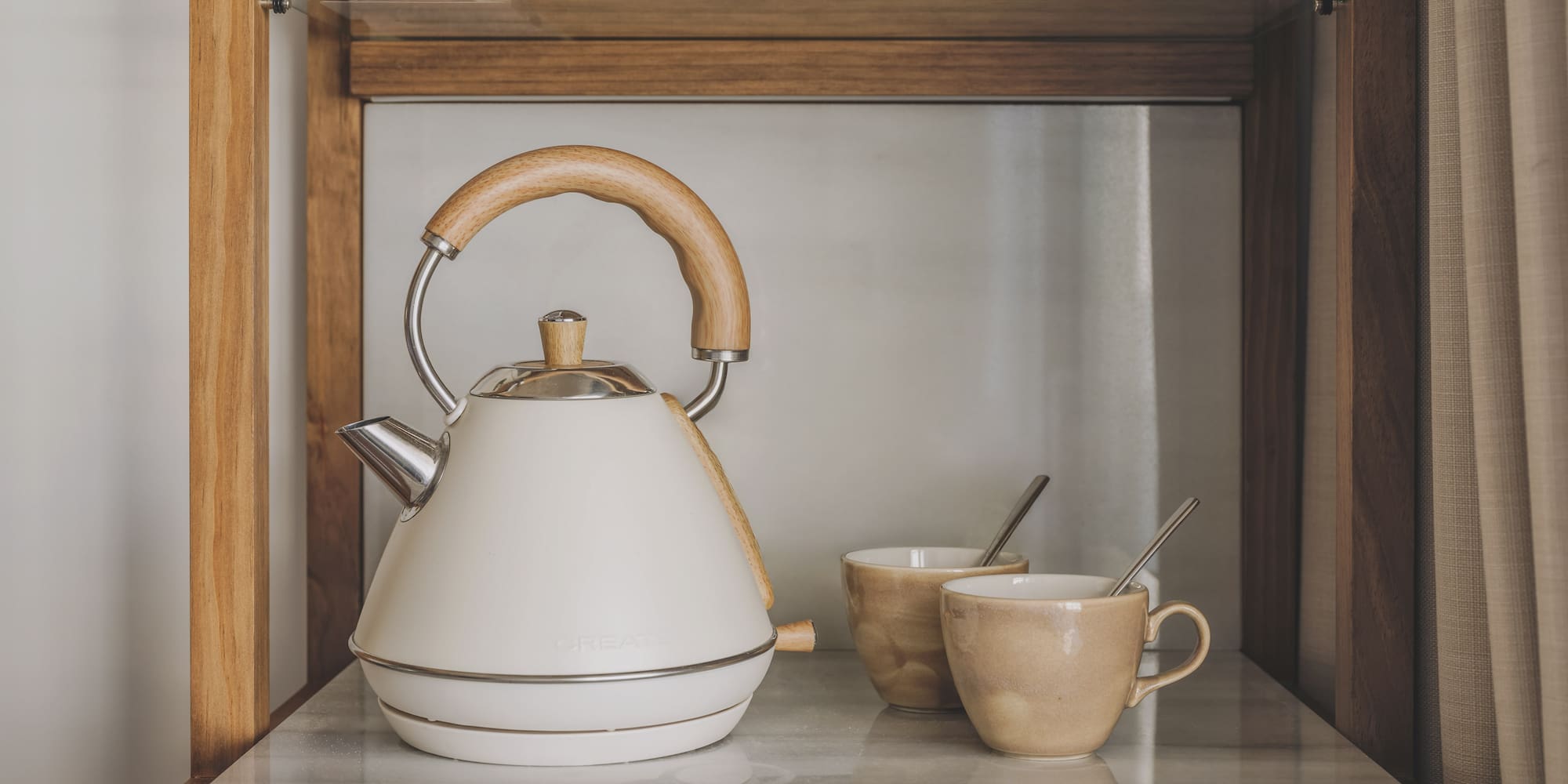 Cream electric kettle, wooden handle, and two mugs on a marble counter.