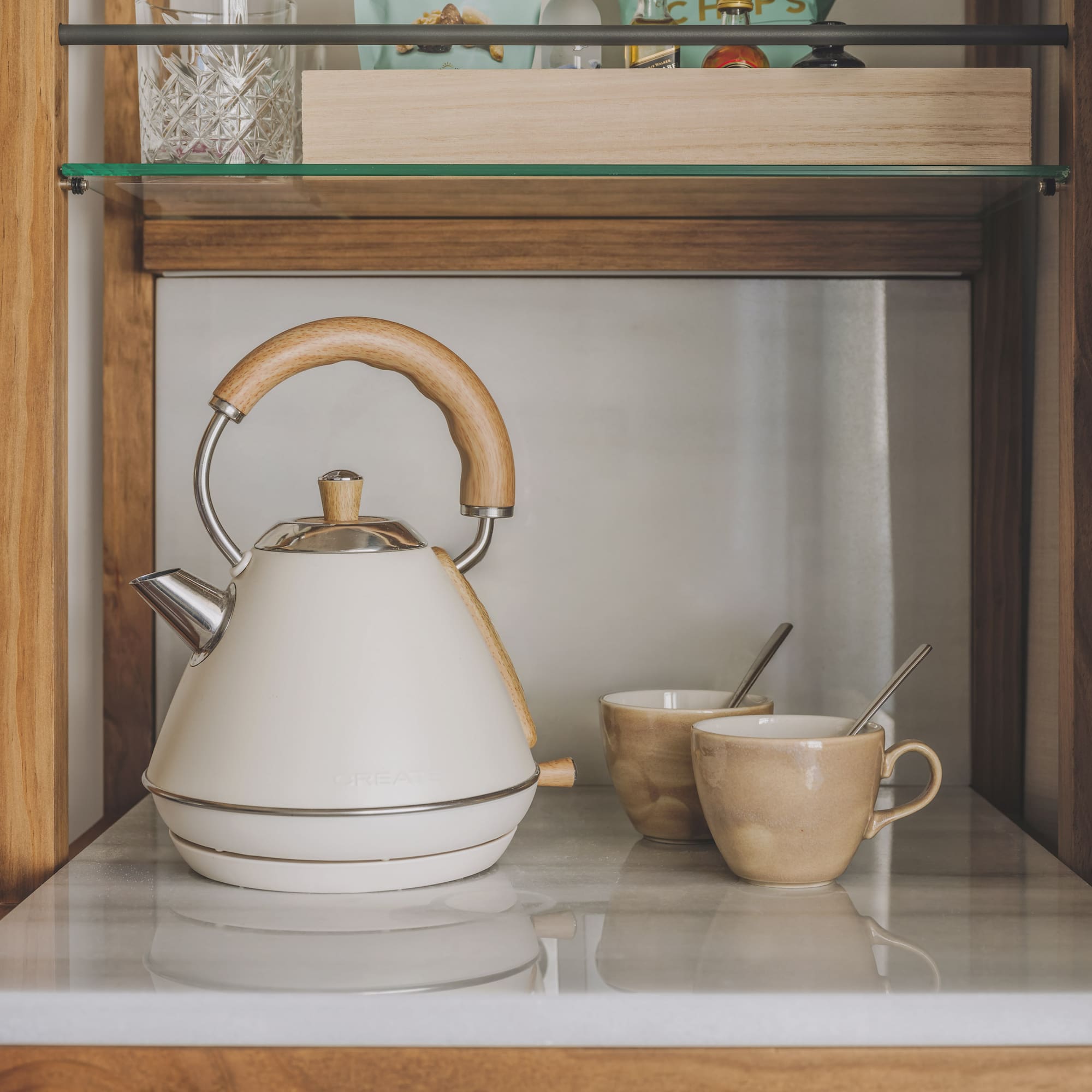 Cream electric kettle with wooden handle, two ceramic tea mugs on marble shelf.