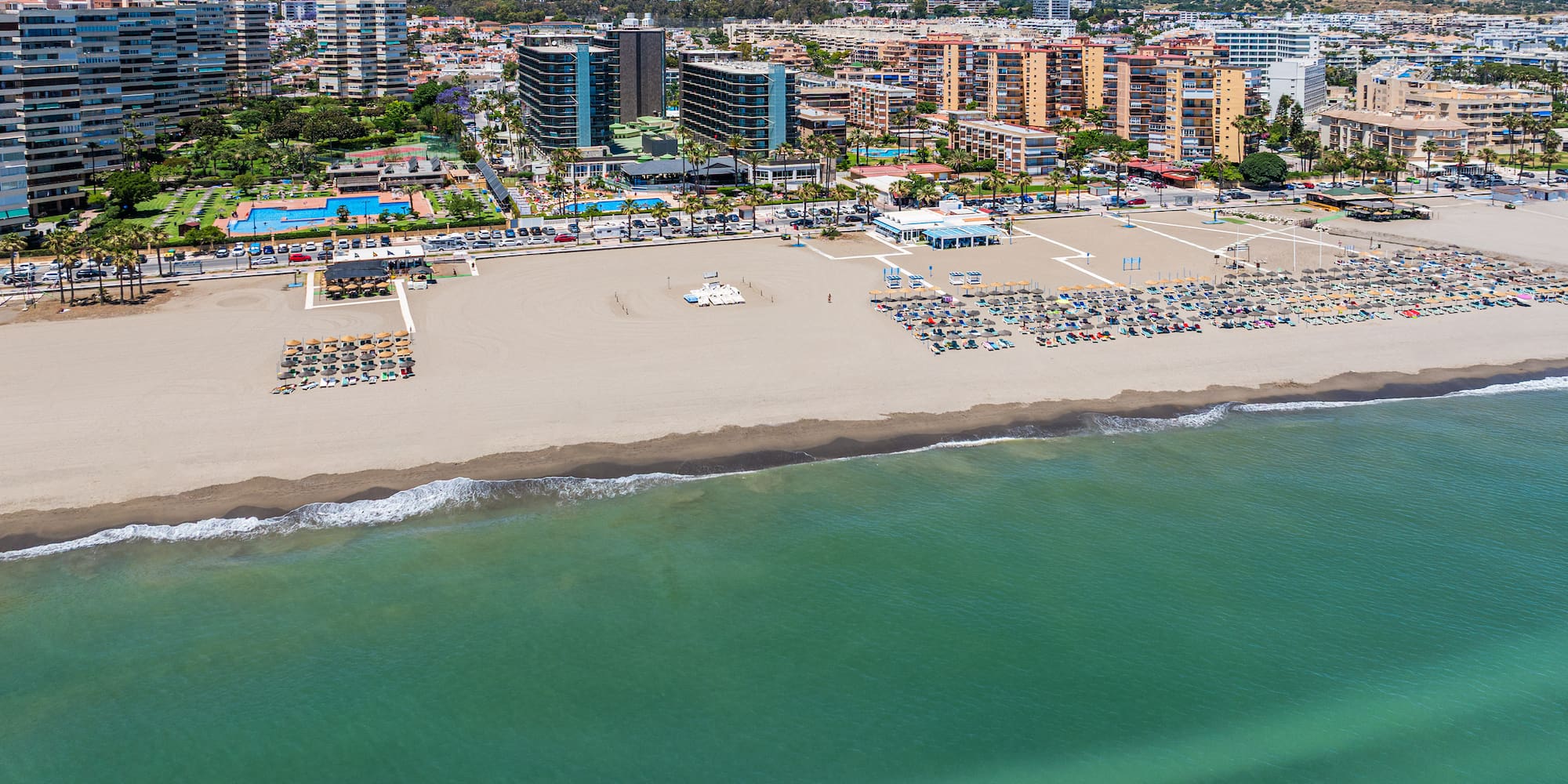 Sunny resort beach with sun loungers, turquoise sea, coastal city, and mountains.