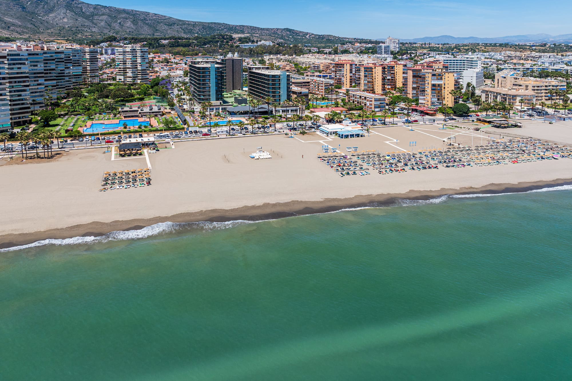 Sunny resort beach with sun loungers, turquoise sea, coastal city, and mountains.