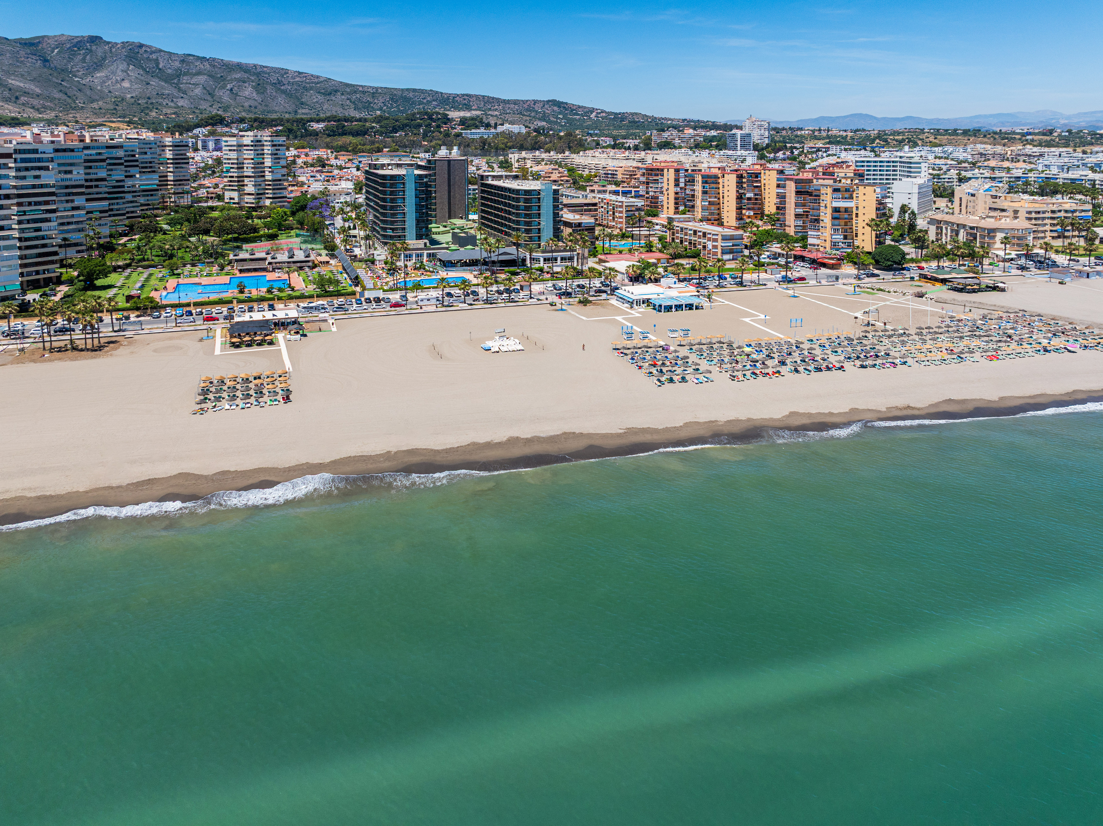 Sunny resort beach with sun loungers, turquoise sea, coastal city, and mountains.