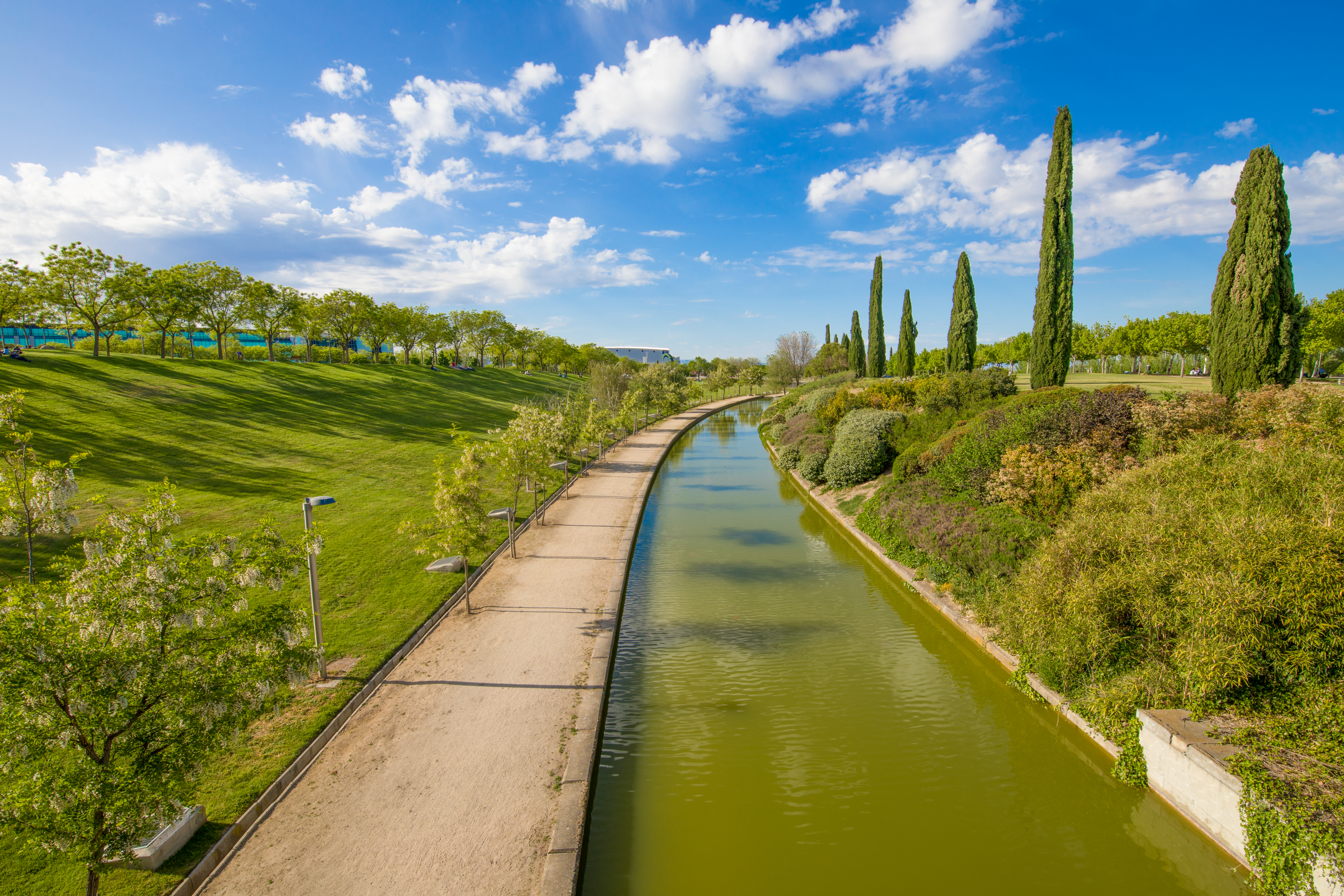 Sunny park with green canal, path, grassy slopes, and tall cypress trees.