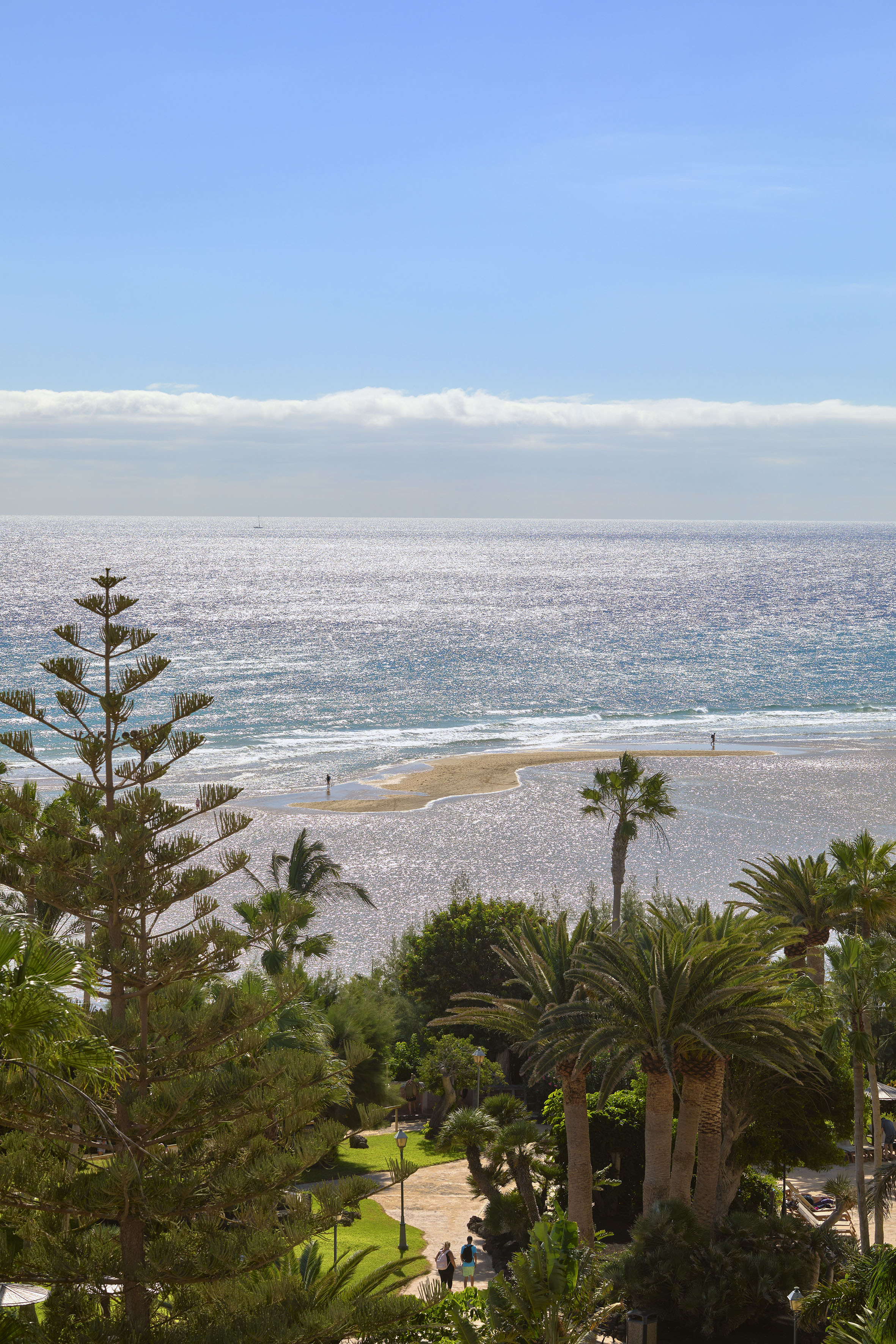 Sunny tropical beach with sparkling sea, sandbar, palm trees, and distant people.