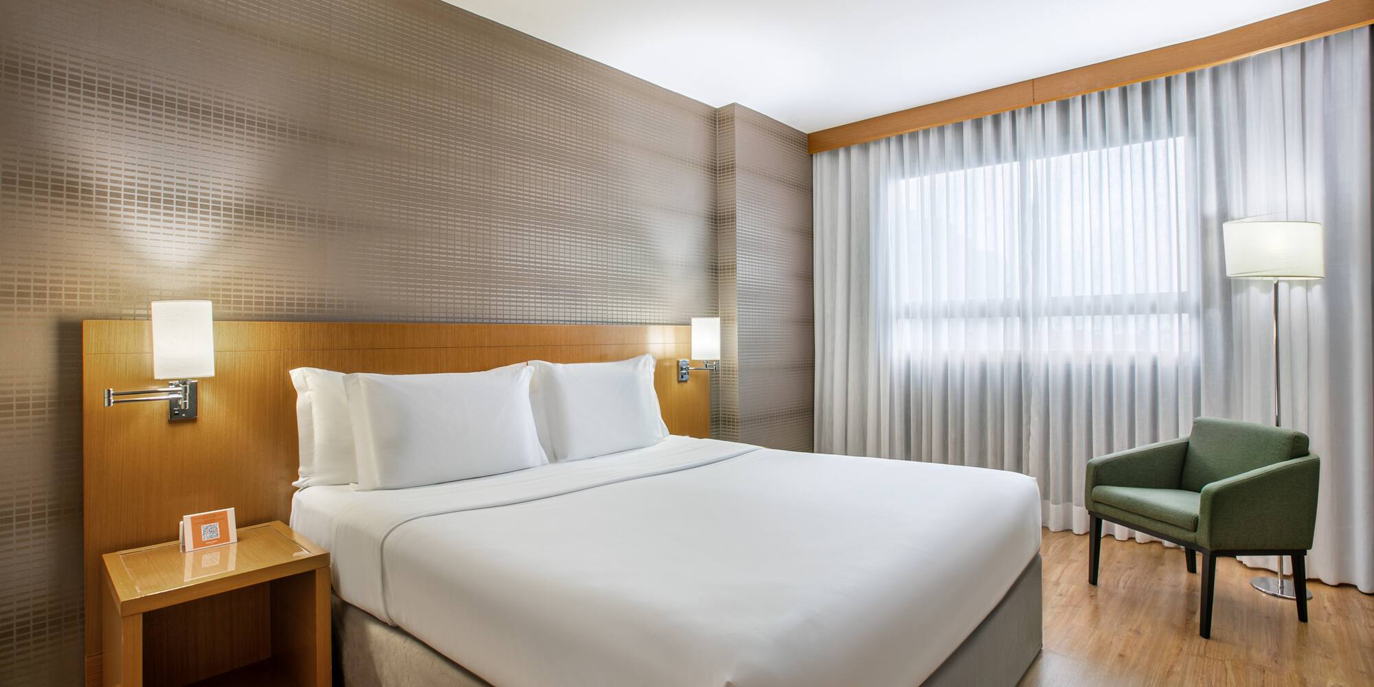 Modern hotel bedroom with king bed, green armchair, and large window.