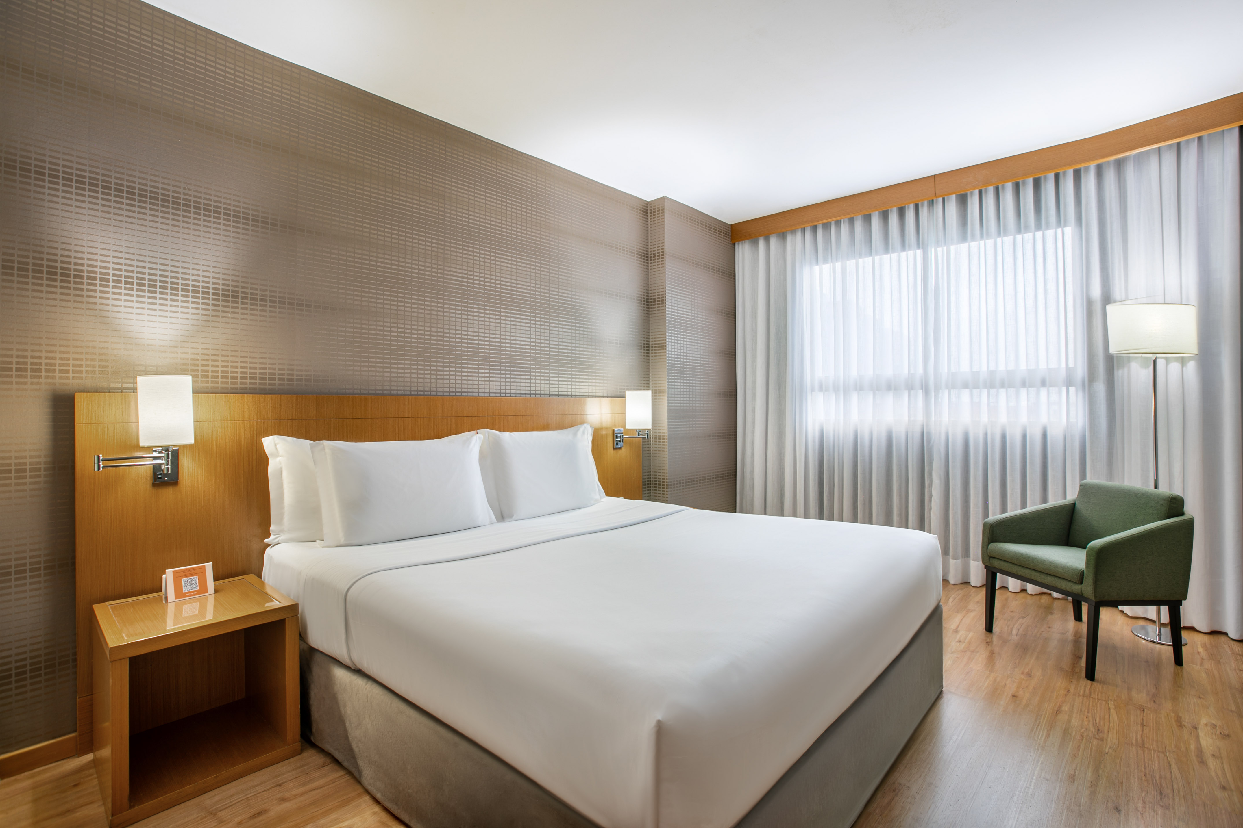 Modern hotel bedroom with king bed, green armchair, and large window.
