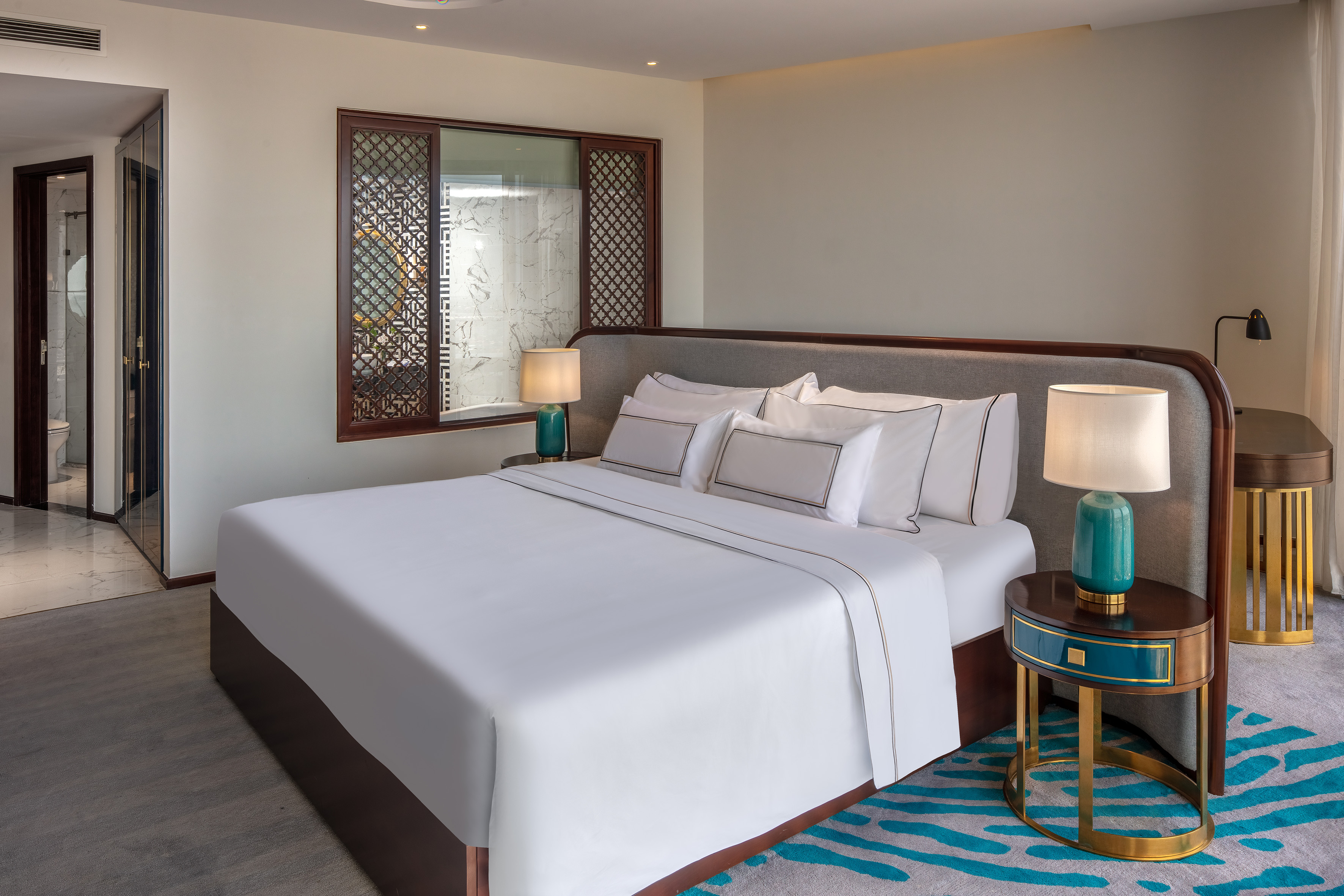 Modern hotel bedroom with king bed, ornate wood screen, and stylish teal decor.