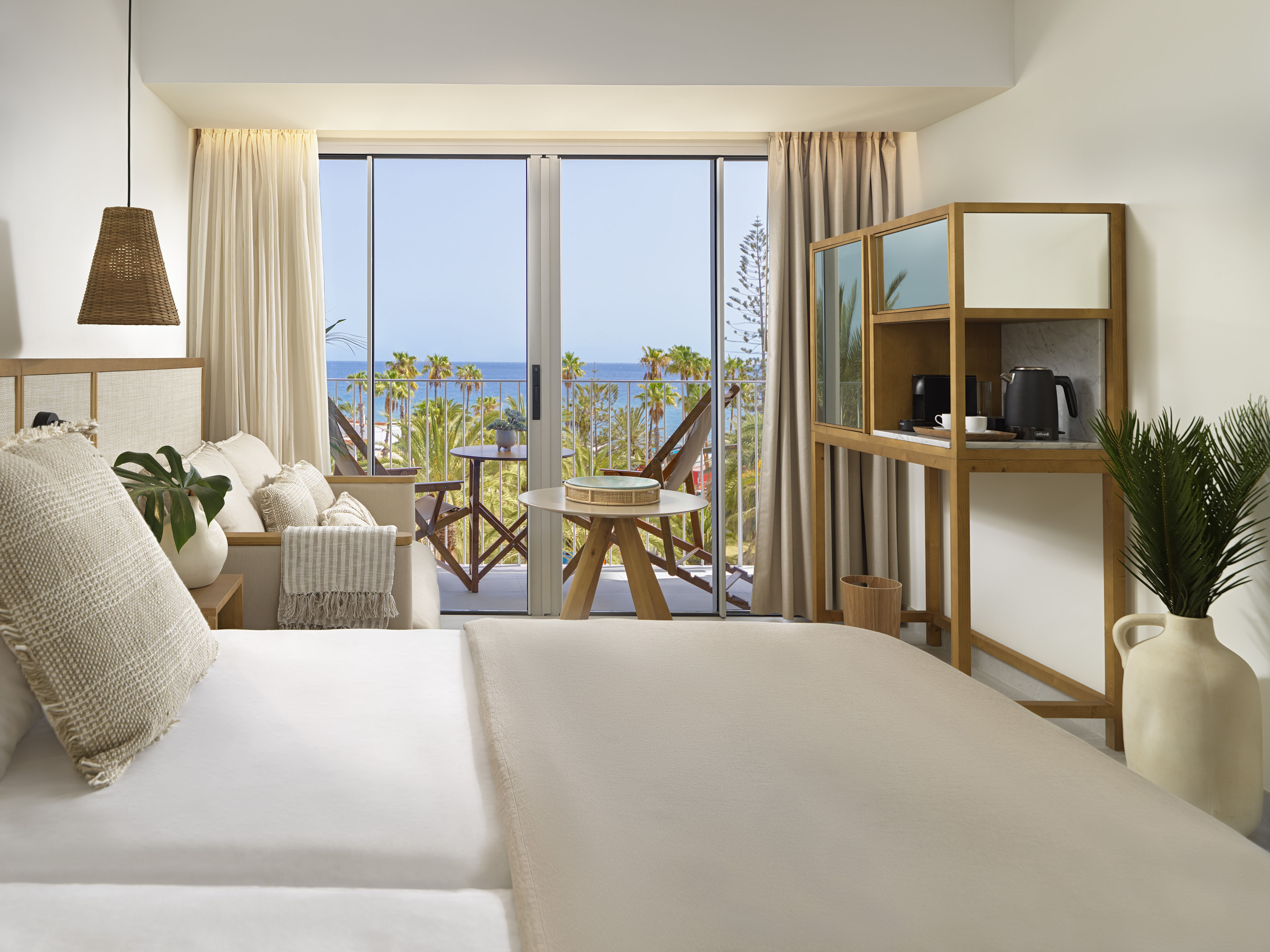Hotel room with bed, balcony ocean view, palm trees, and coffee station.