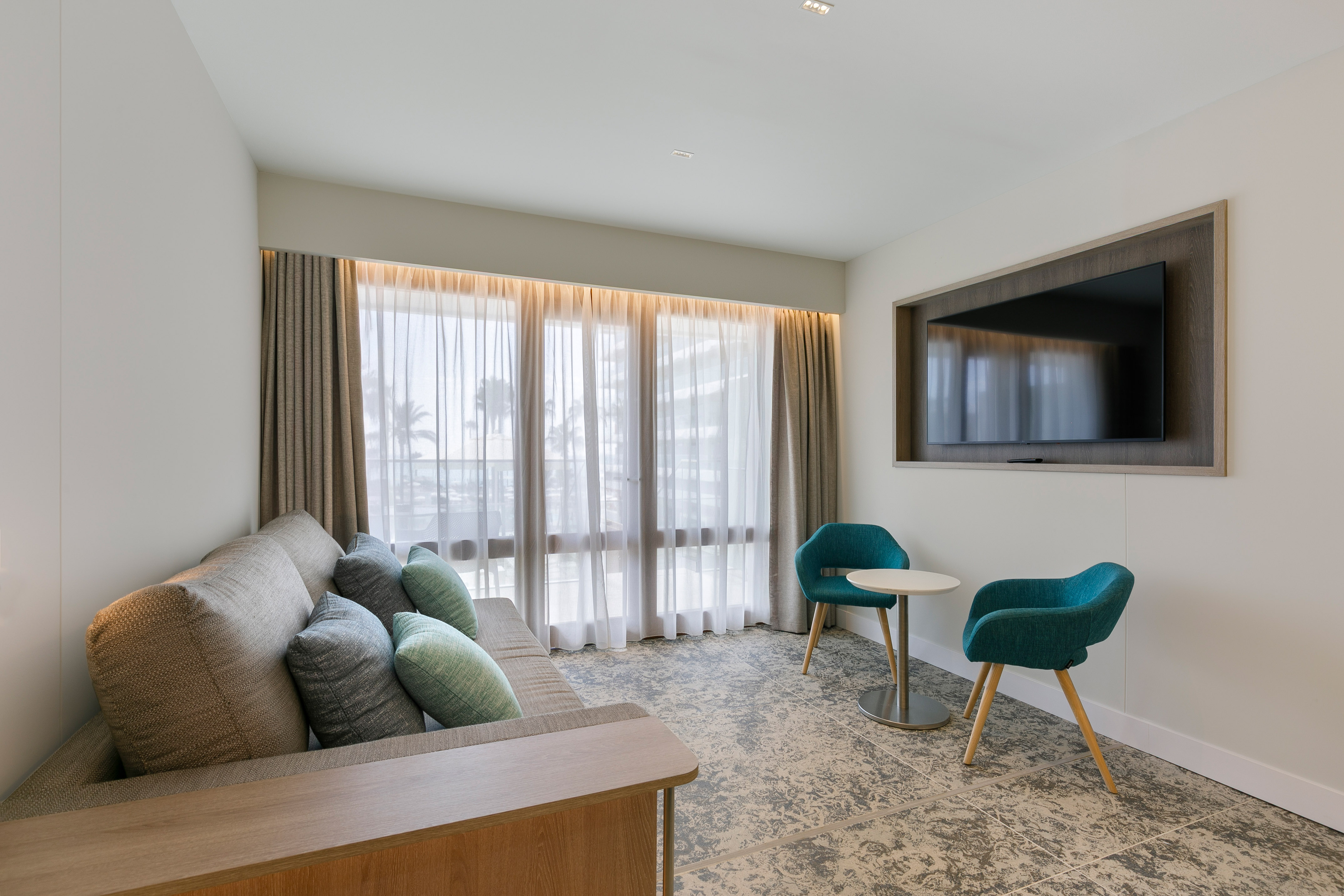 Modern hotel suite living room with sofa, wall TV, and teal chairs.