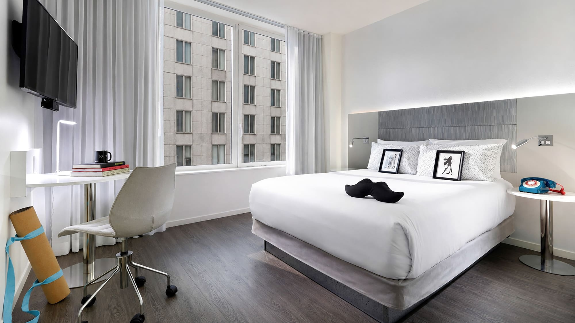Stylish hotel bedroom with king bed, work desk, yoga mat, and city window view.