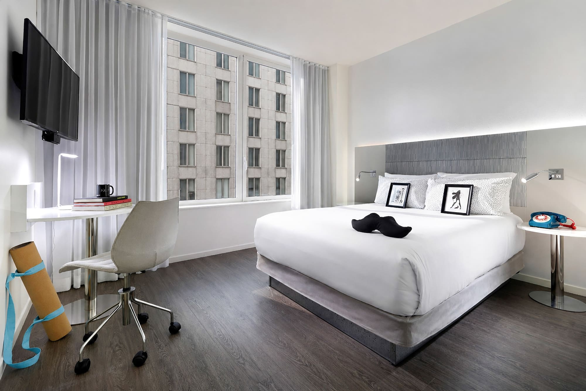 Stylish hotel bedroom with king bed, work desk, yoga mat, and city window view.