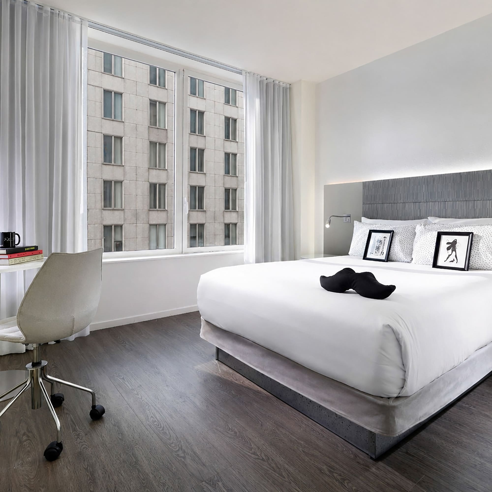 Stylish hotel bedroom with king bed, work desk, yoga mat, and city window view.