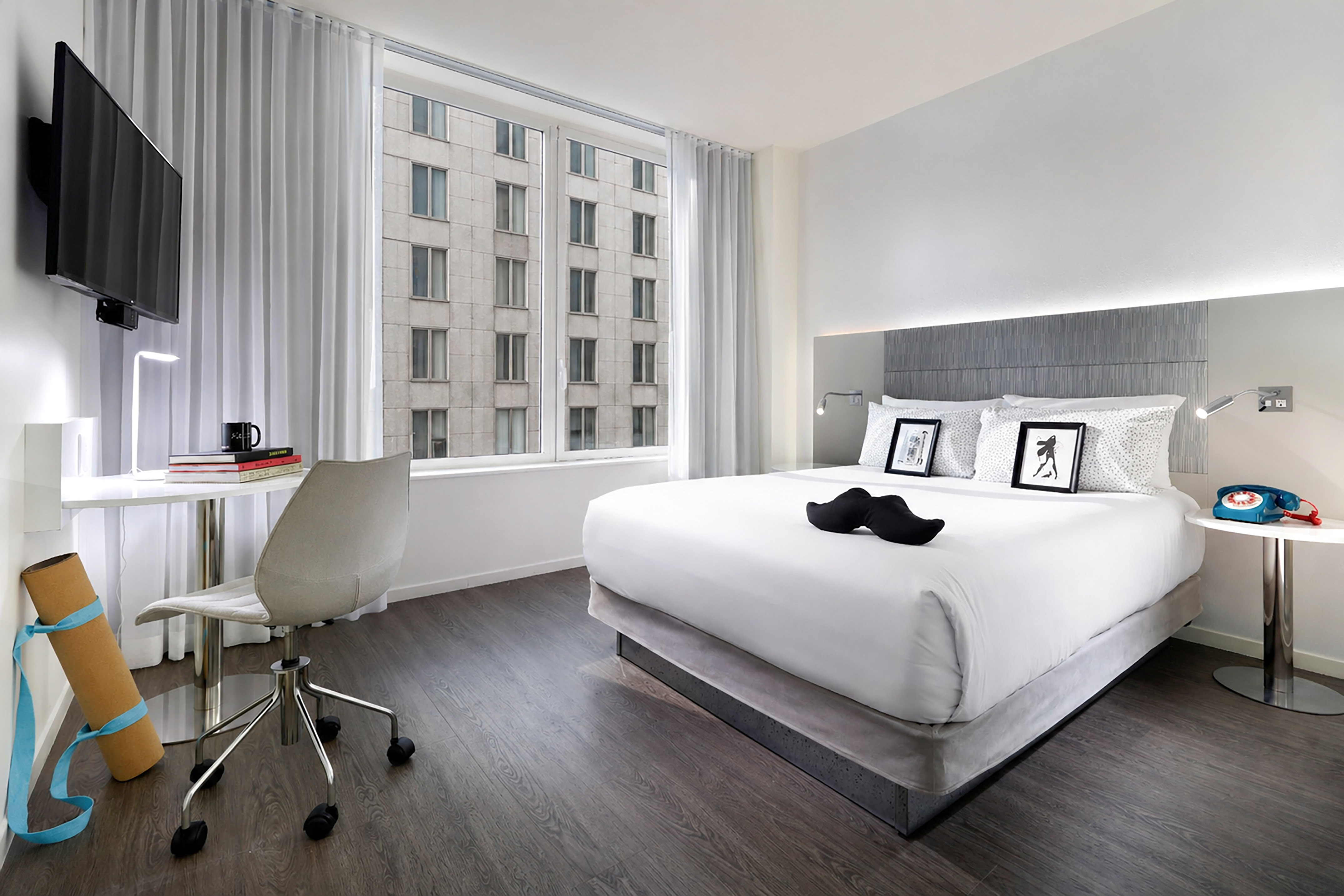 Stylish hotel bedroom with king bed, work desk, yoga mat, and city window view.