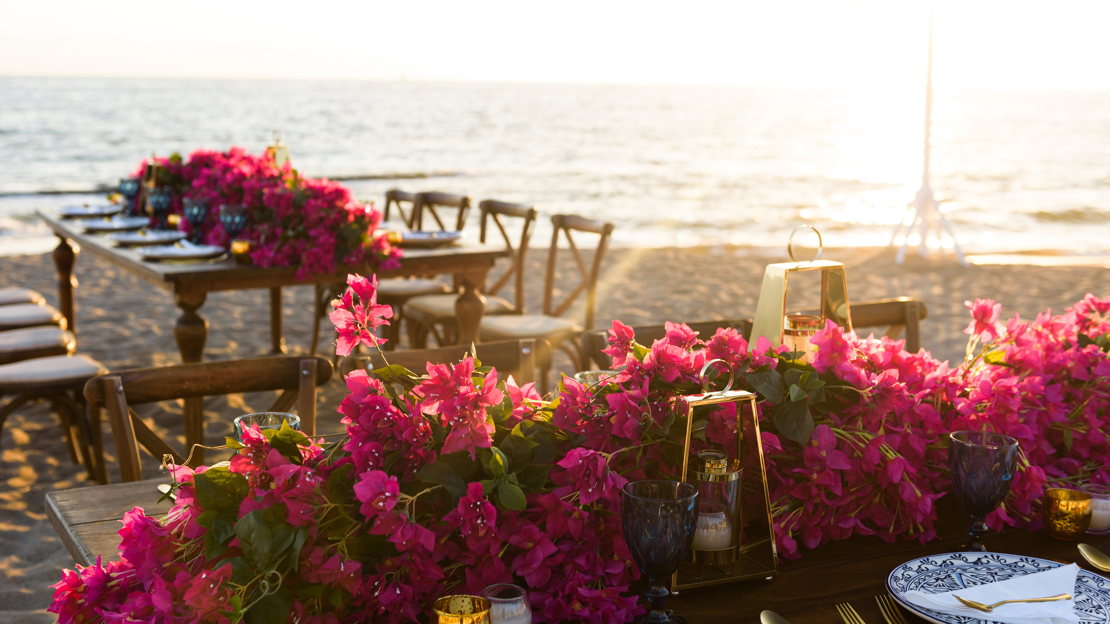 Beach dining table with vibrant pink bougainvillea, gold lanterns, sunset ocean.