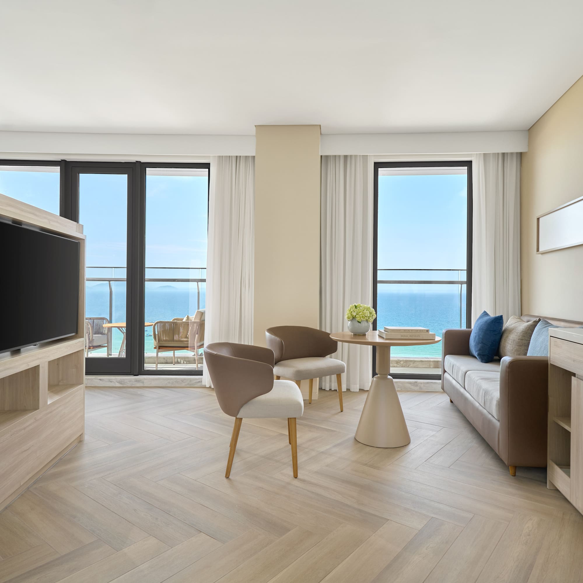Modern hotel suite living room with ocean view, herringbone floor, and balcony.