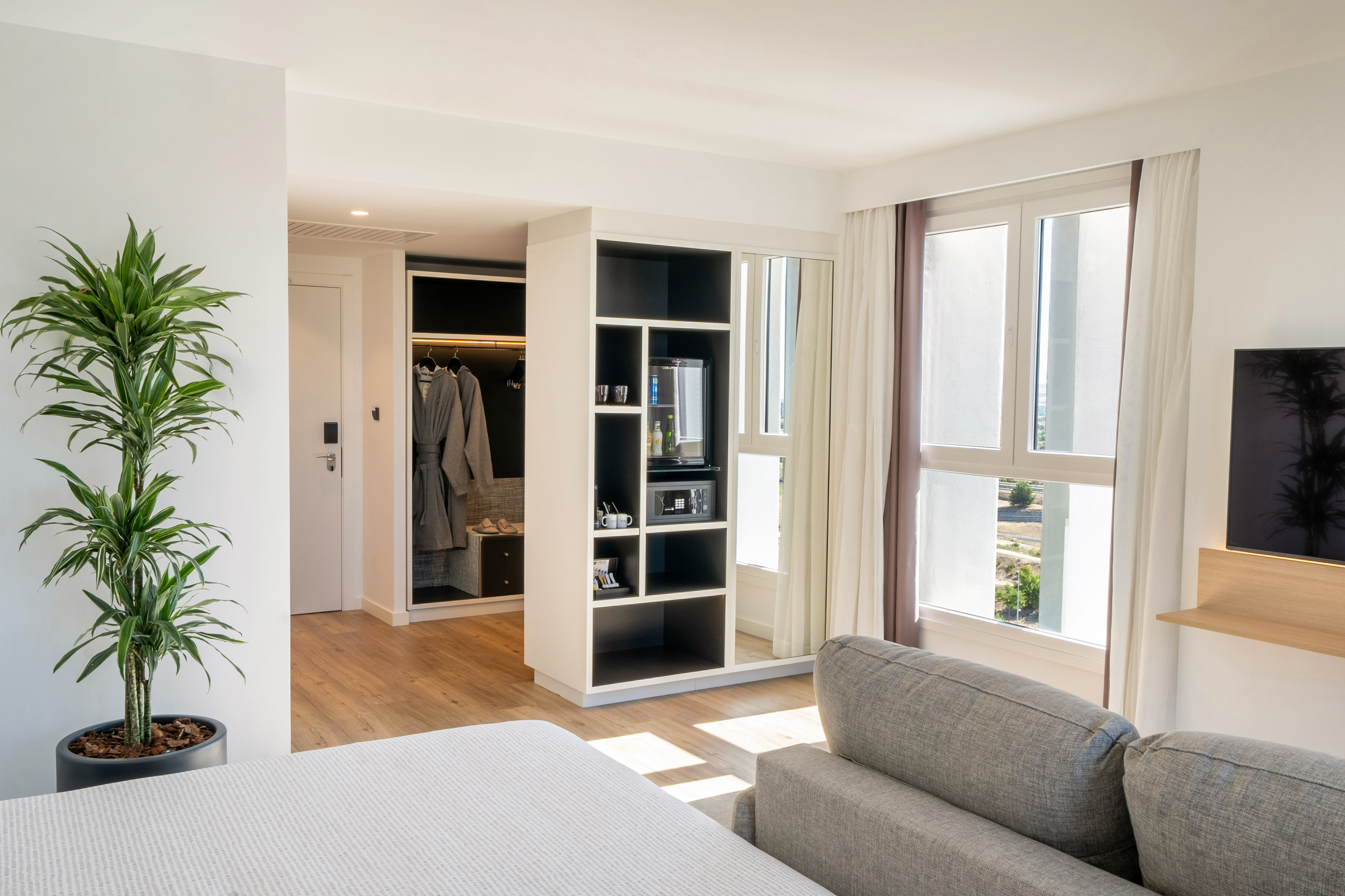 Modern hotel suite with bed, sofa, green plant, minibar, and large window.