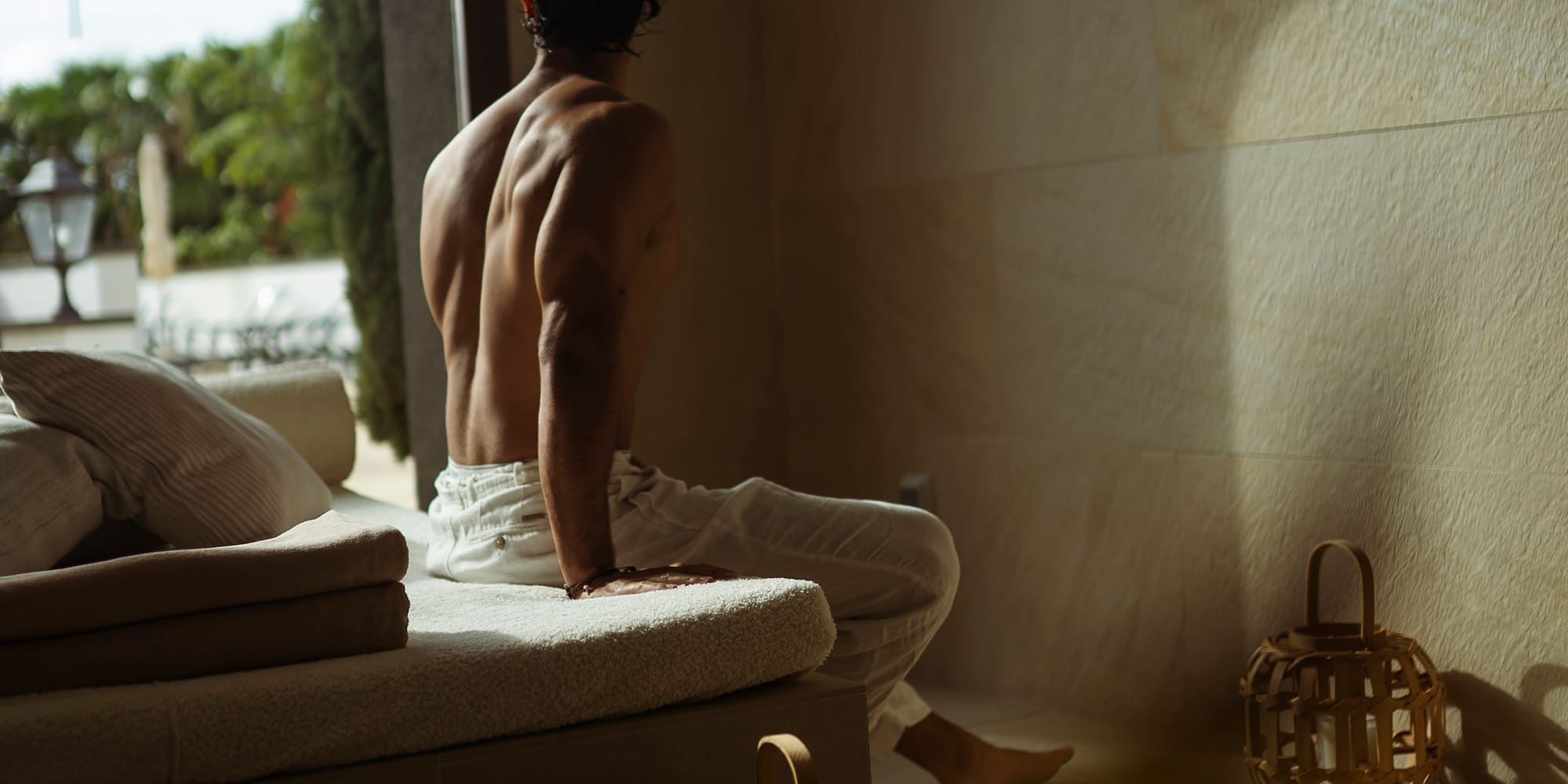 Shirtless man with muscular back relaxing on a bench, looking out arched doorway.