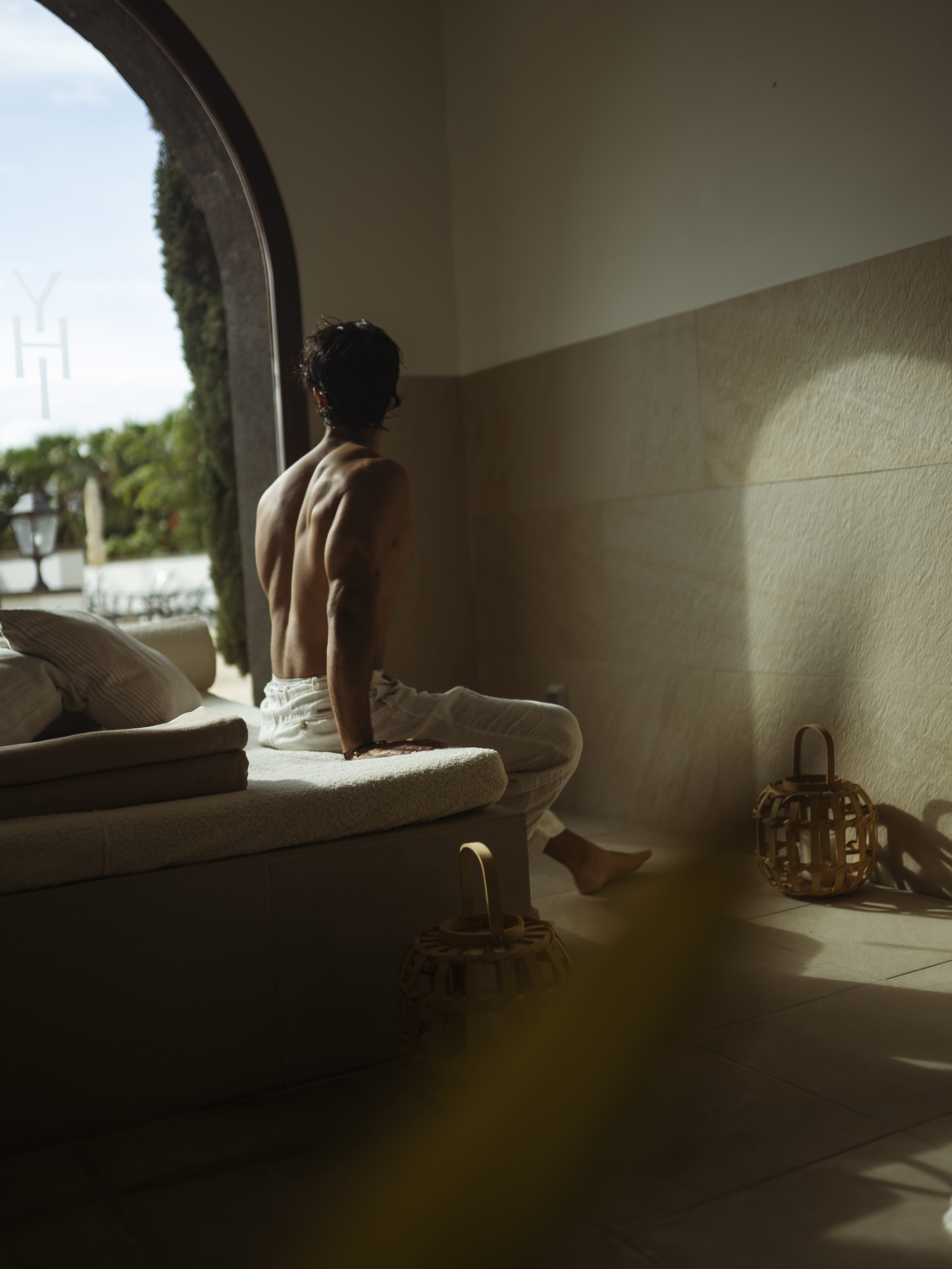 Shirtless man with muscular back relaxing on a bench, looking out arched doorway.