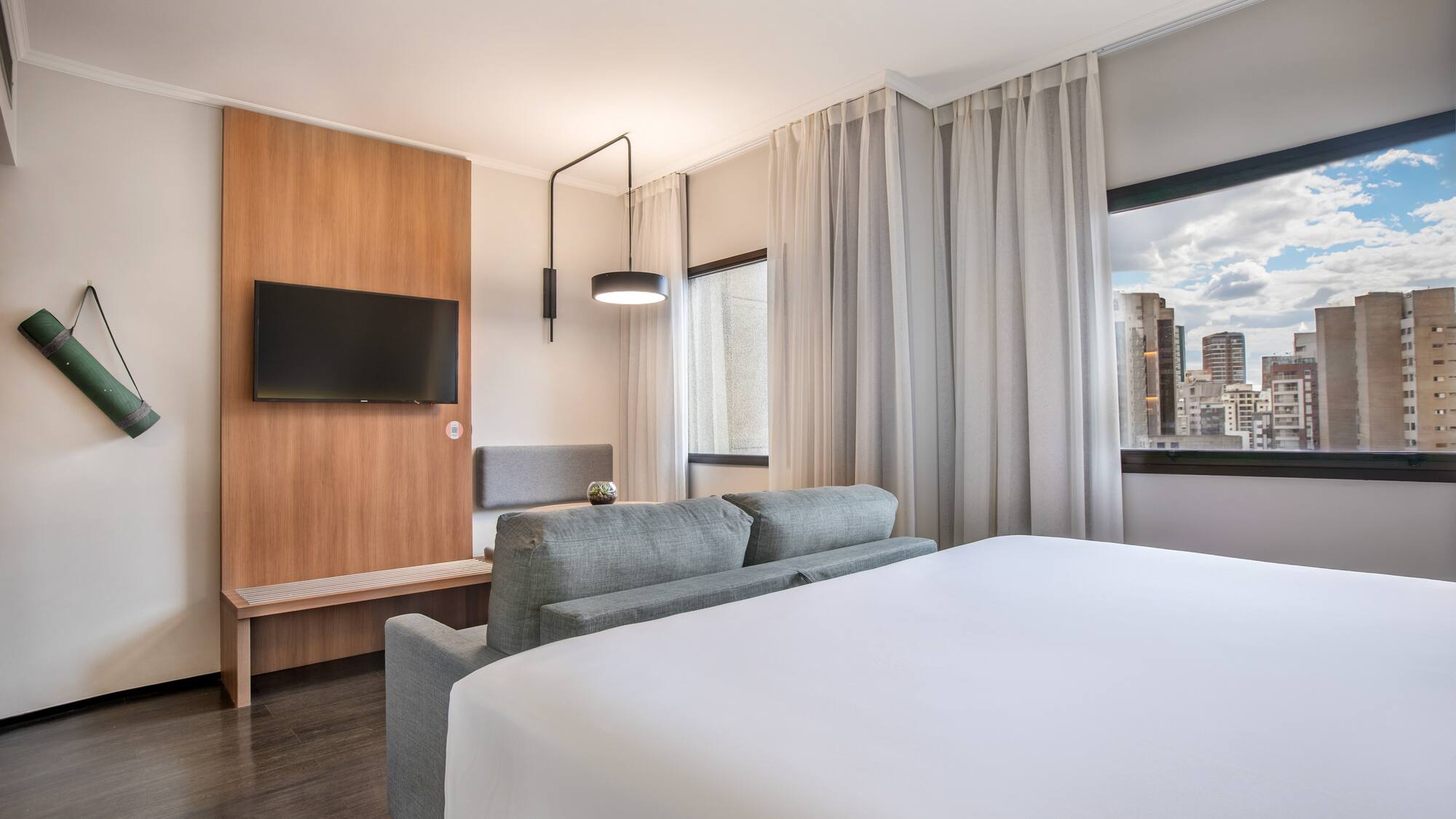 Modern hotel suite with bed, sofa, TV, yoga mat, and city skyline view.