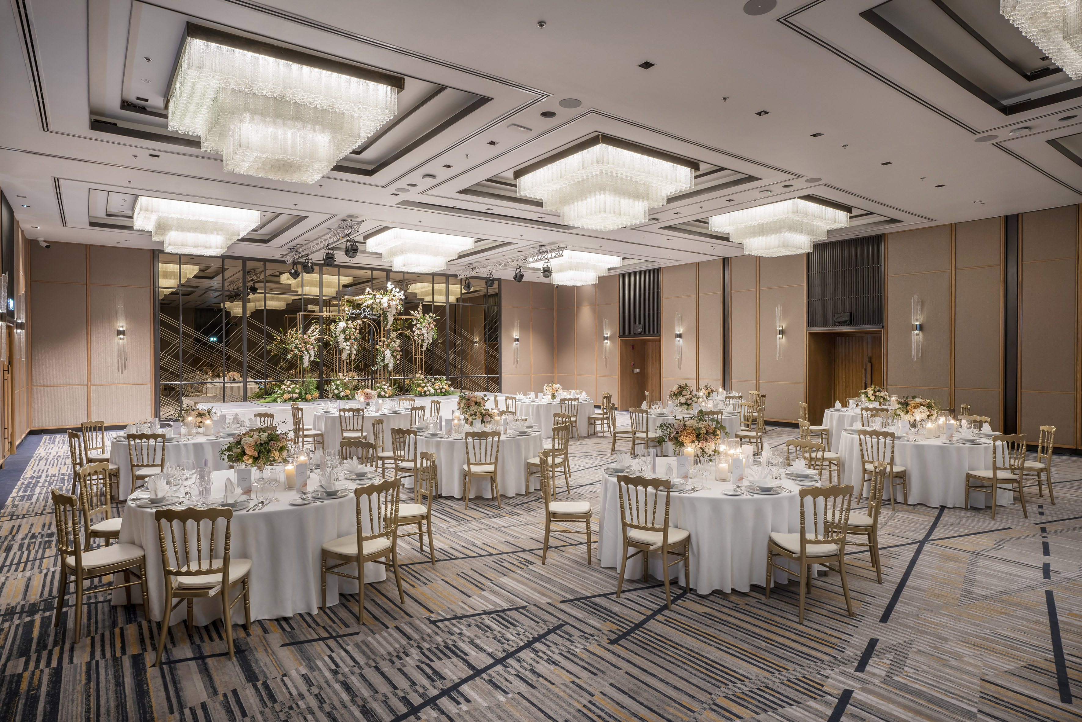 Elegant ballroom event setup with round tables, gold chairs, floral decor, and chandeliers.