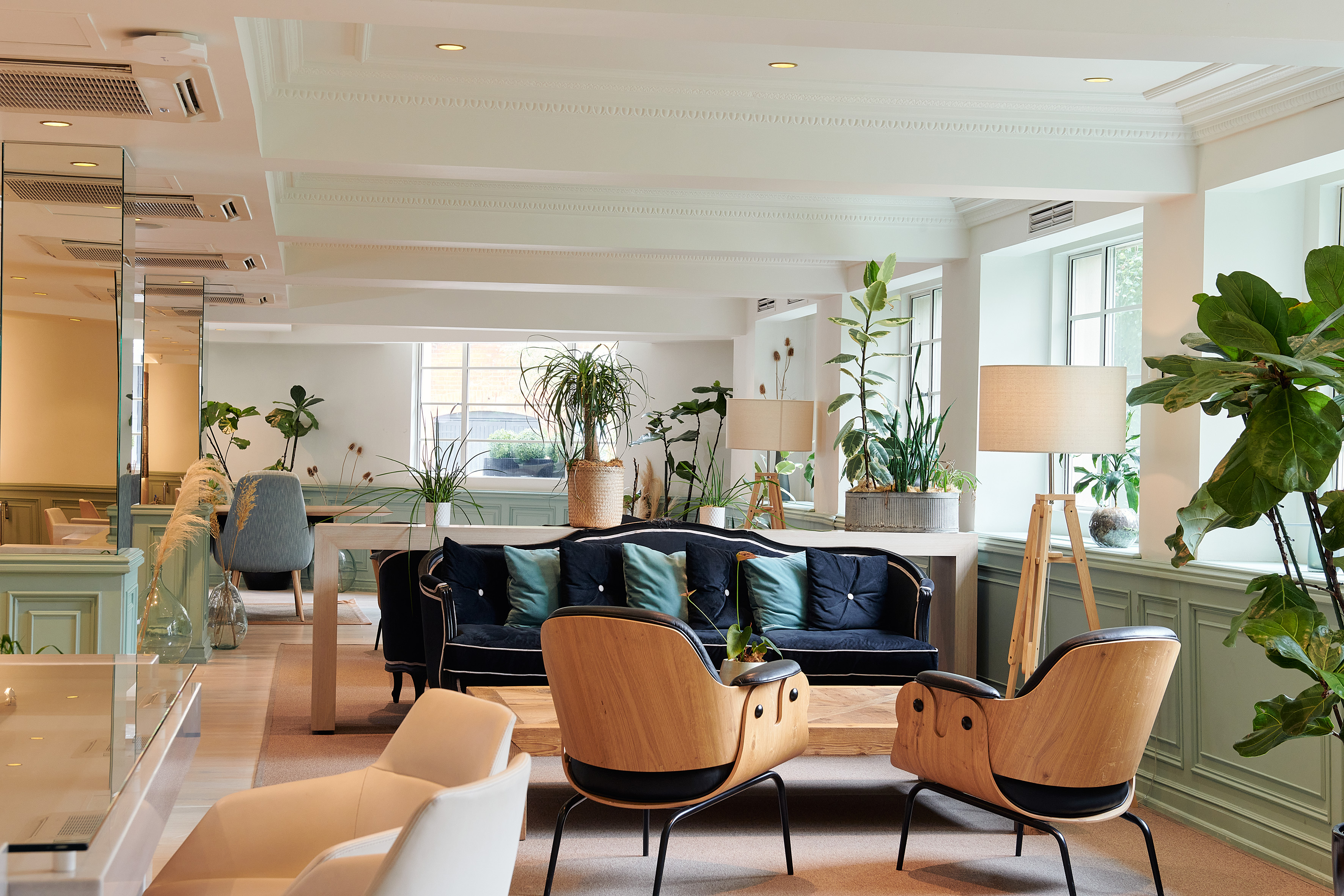 Modern hotel lounge with dark sofa, wooden chairs, ample greenery.